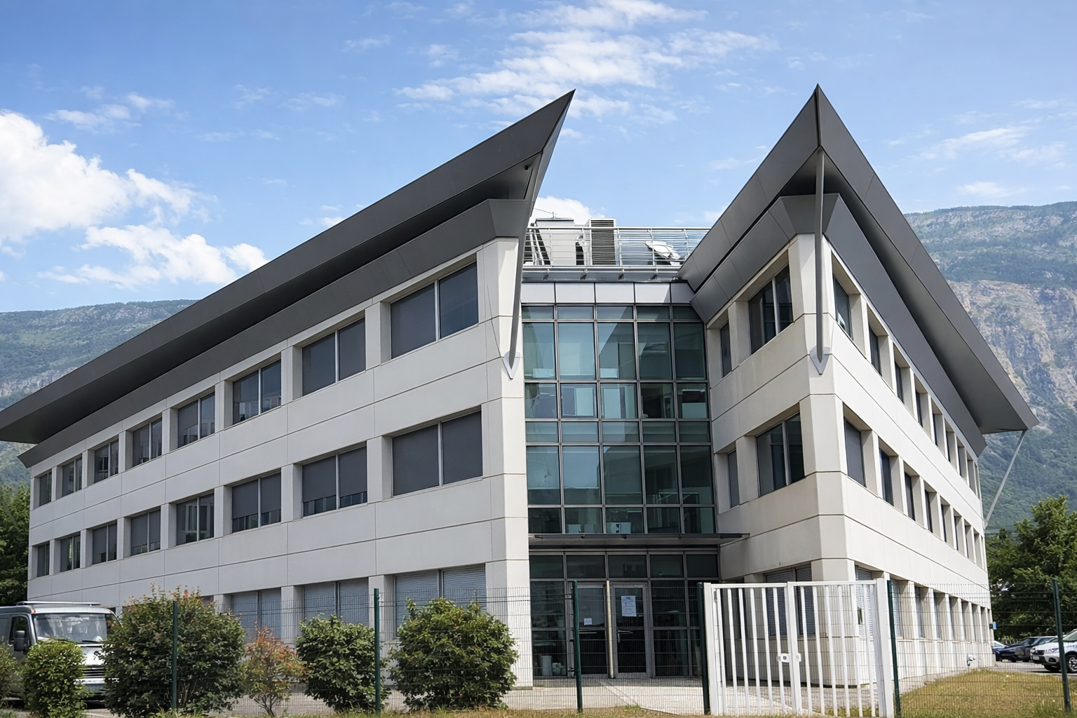 Bâtiment moderne à plusieurs étages avec architecture angulaire, grandes fenêtres et toit incliné, en arrière-plan des montagnes.