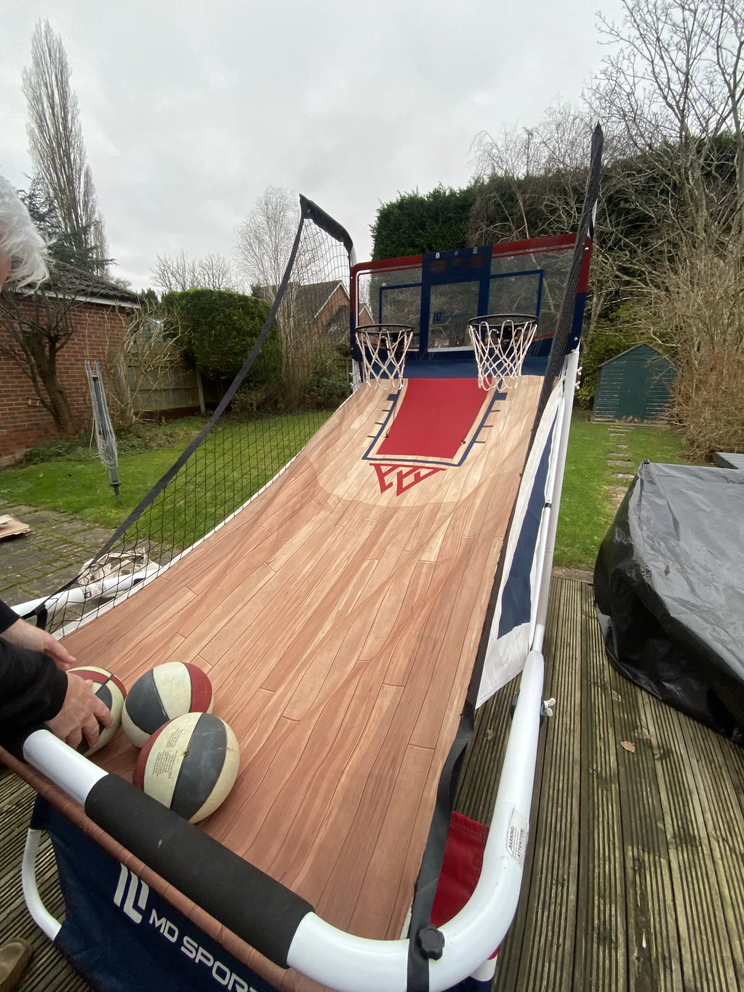 A basketball arcade game with a wooden ramp and two hoops, set up outdoors on a wooden deck in a backyard.