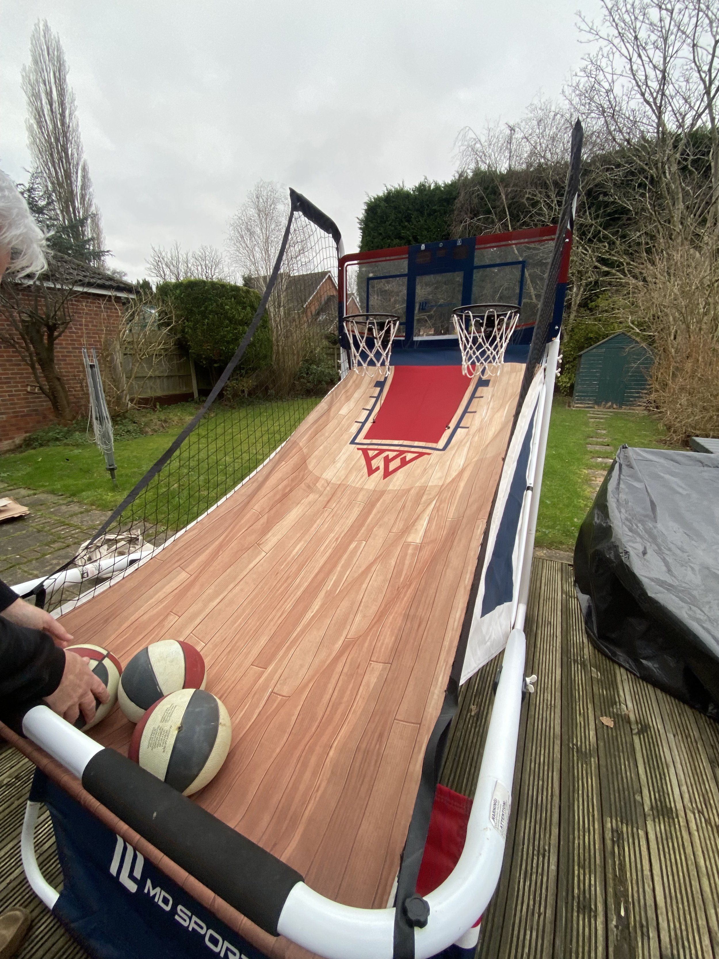 A backyard basketball arcade game with a wooden sliding surface, two basketball hoops, and a net side barrier, set up on a wooden deck outside.