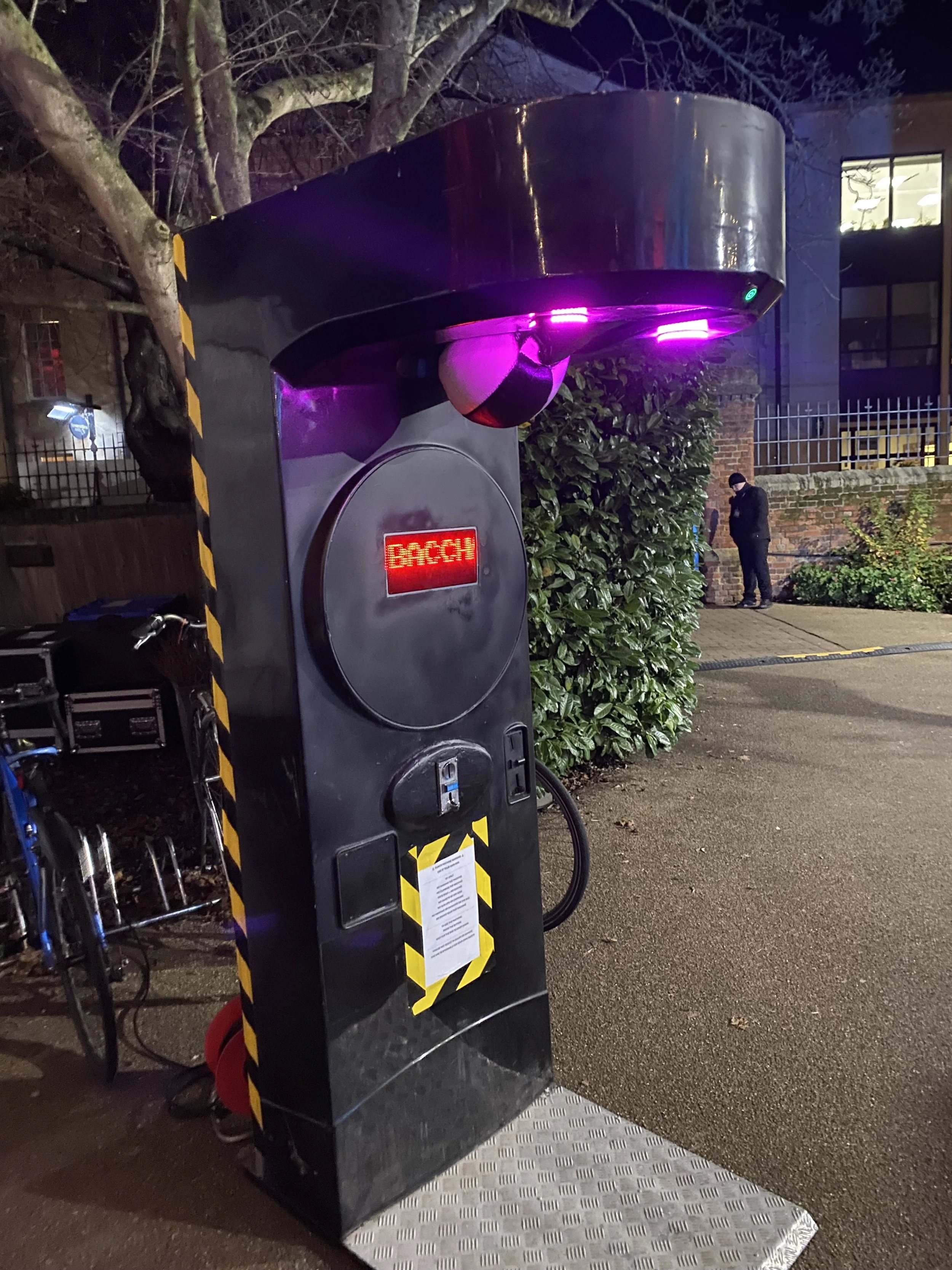 Electric vehicle charging station with a screen displaying 'BACCH' and purple lights, located outdoors at night with a person and some greenery in the background.