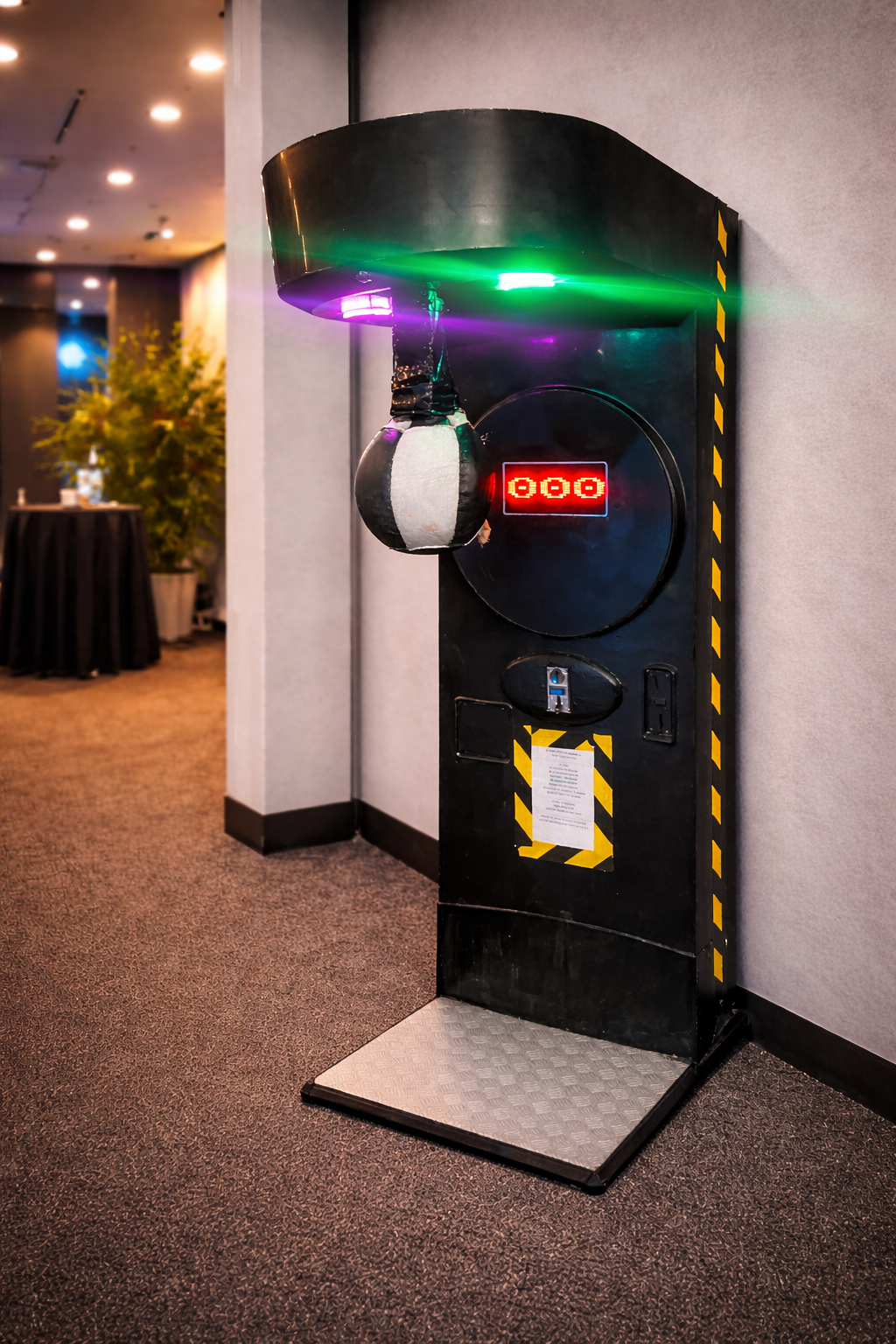 A black punching machine with a digit display showing three red zeros, green and purple lights at the top, and a padded punching ball hanging from above, situated in an indoor setting with a carpeted floor and a large potted plant in the background.