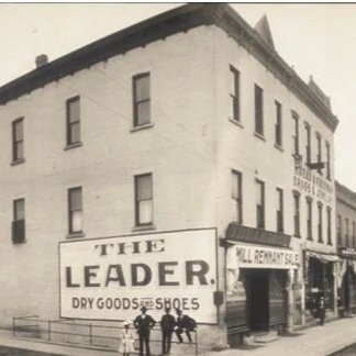 Corner view of store. Giant sign on building taking up a floor that says The LEADER dry goods and shoes.