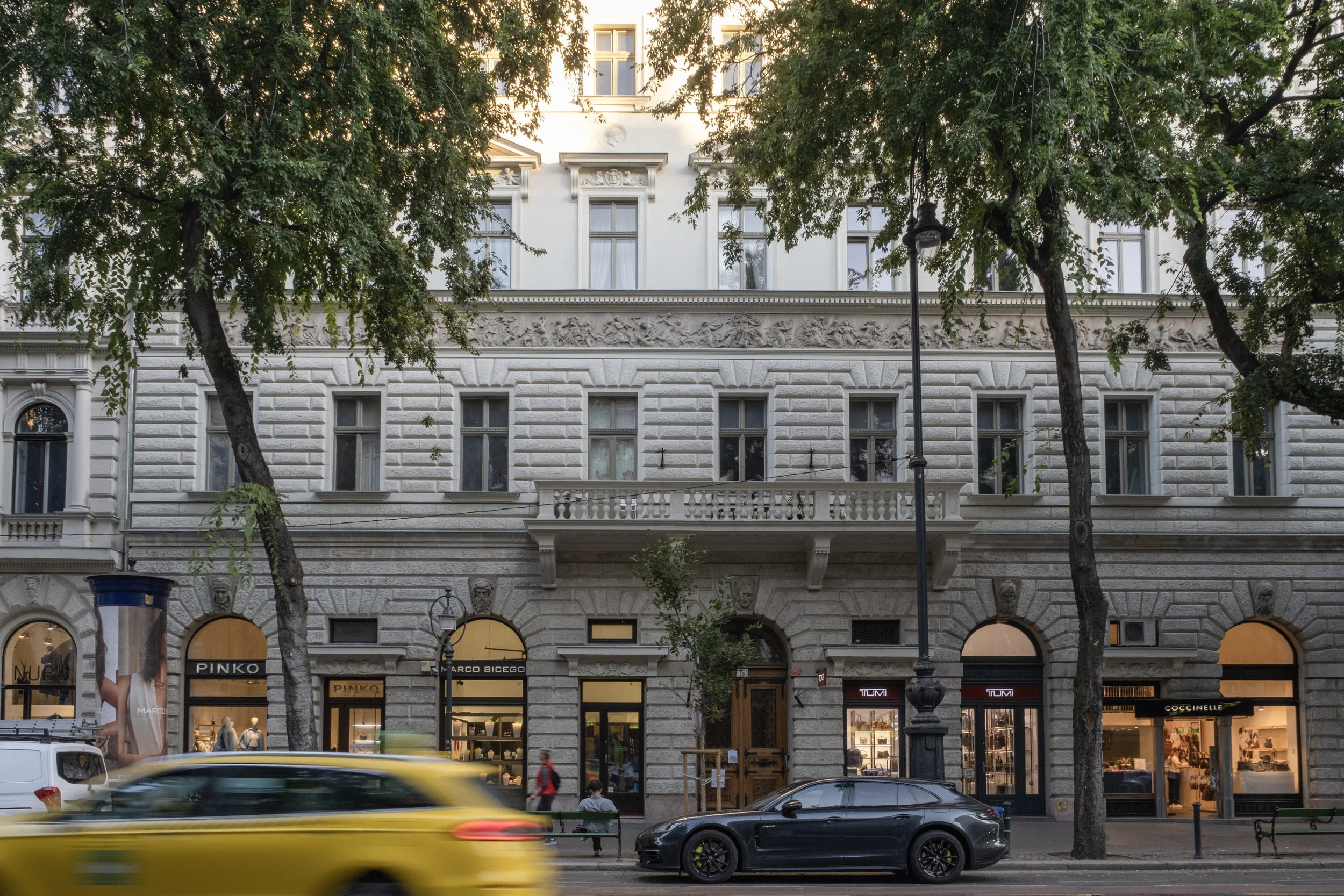 City street scene with a white ornate building in the background, trees partially obscuring the facade, and moving yellow and black cars passing by in the foreground.