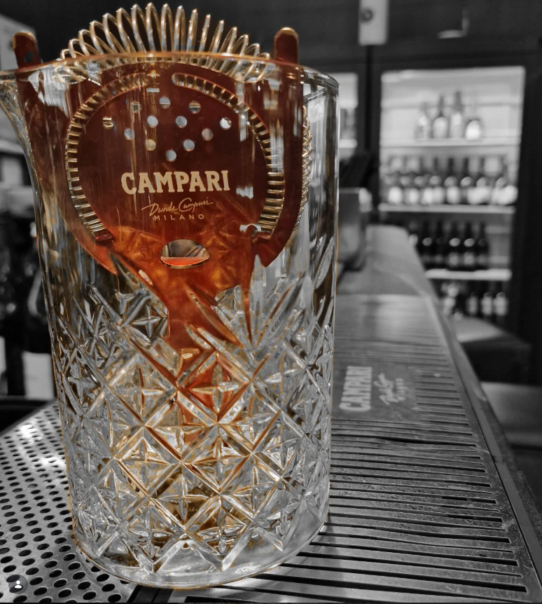 Un bicchiere di cristallo decorato con un disegno inciso, con al suo interno una bustina di cioccolato al liquore Campari, in un bancone di bar.