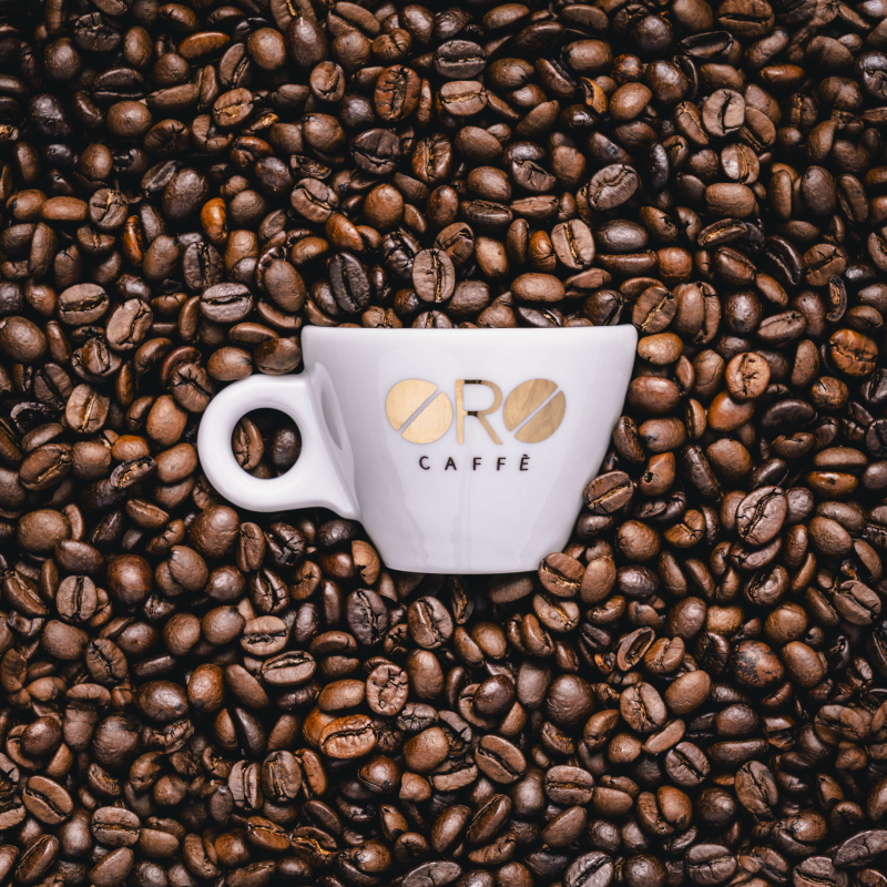 Tazza bianca con il logo 'ORO CAFFÈ' appoggiata su una superficie ricoperta di chicchi di caffè tostato.