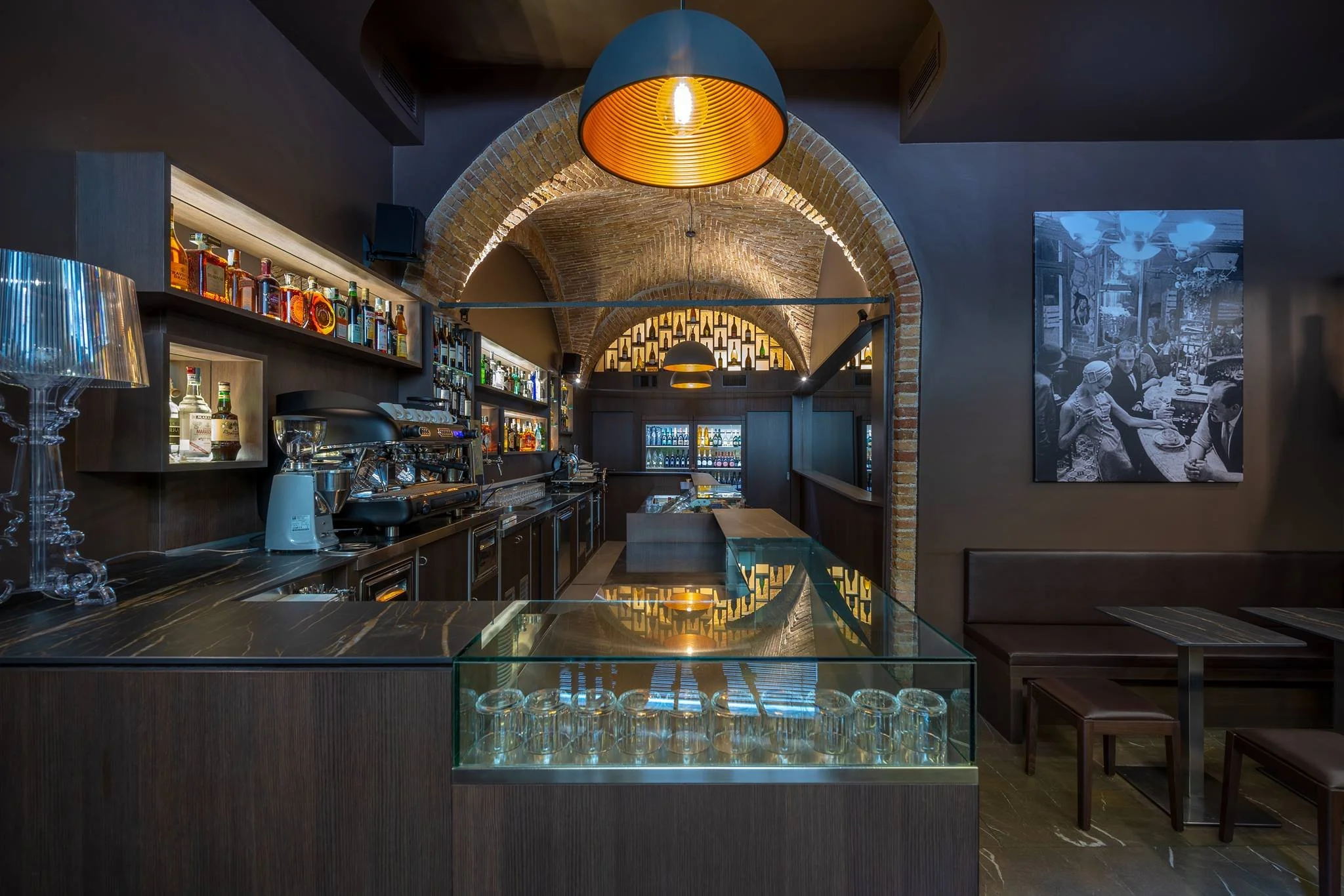 Interno di un bar con archi in mattoni, scaffali con bottiglie di alcol, bancone con macchina da caffè, tavoli e sedie in legno, e un quadro in bianco e nero alle pareti.