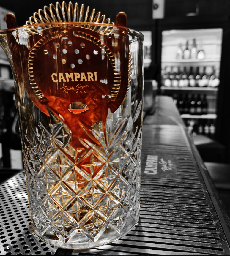 Un bicchiere di cristallo decorato con motivo a croce contenente un liquido rosso nelle mani di qualcuno, con uno sfondo di supermercato con scaffali di bottiglie.