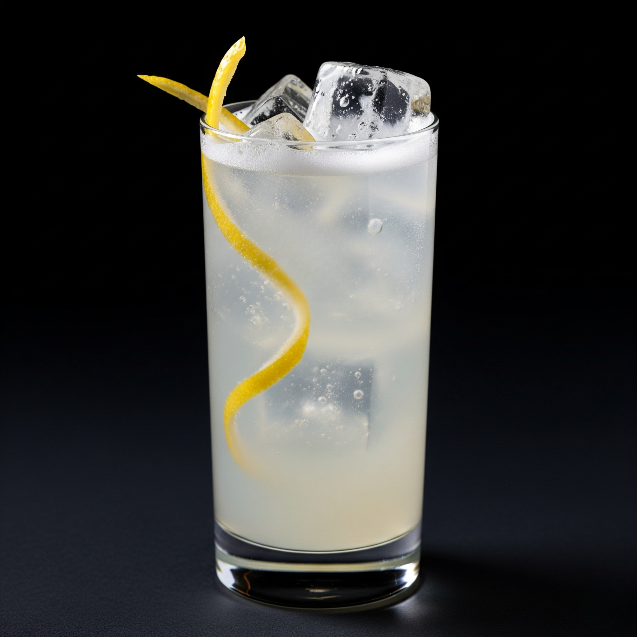 Bicchiere alto con cocktail al limone, con due cubetti di ghiaccio e una spirale di scorza di limone, su sfondo nero.