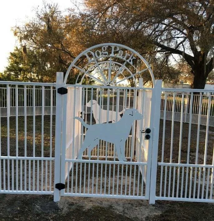 White metal gate with dog silhouette decorations and a sign above that reads 'Dog Trot' in a park with trees in the background.
