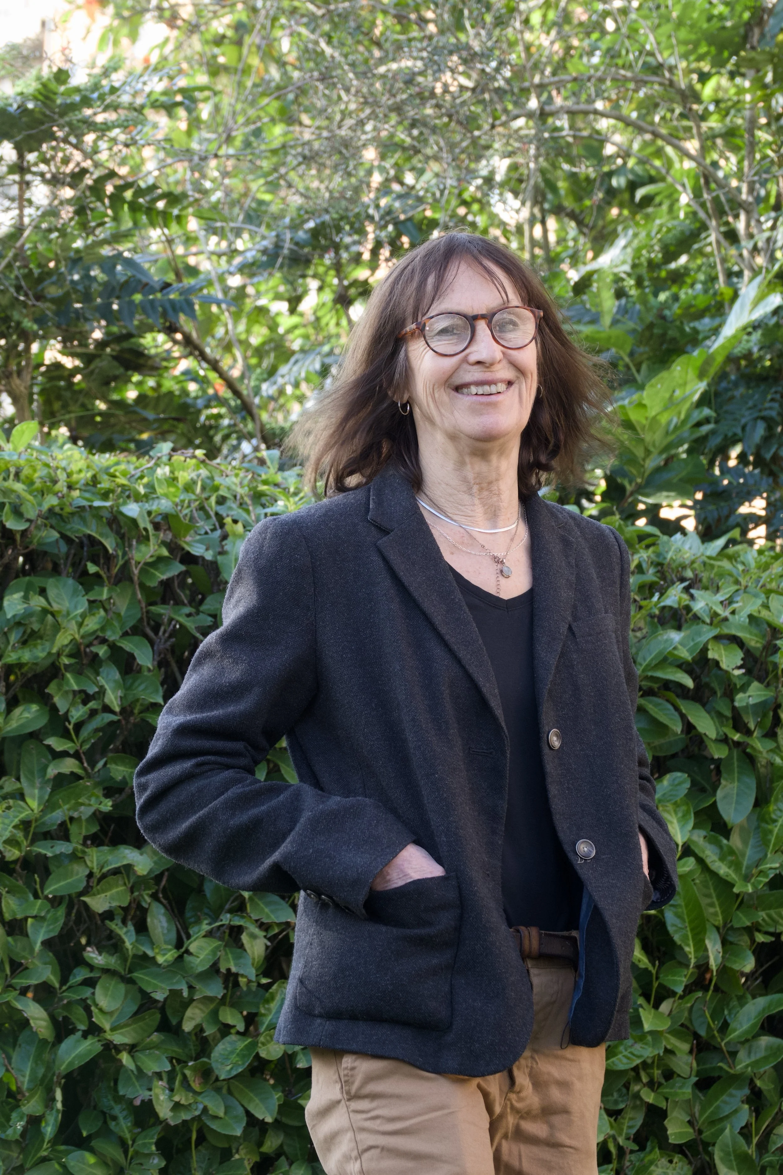 A smiling woman with glasses, wearing a dark blazer and a black top, standing outdoors in front of green foliage.