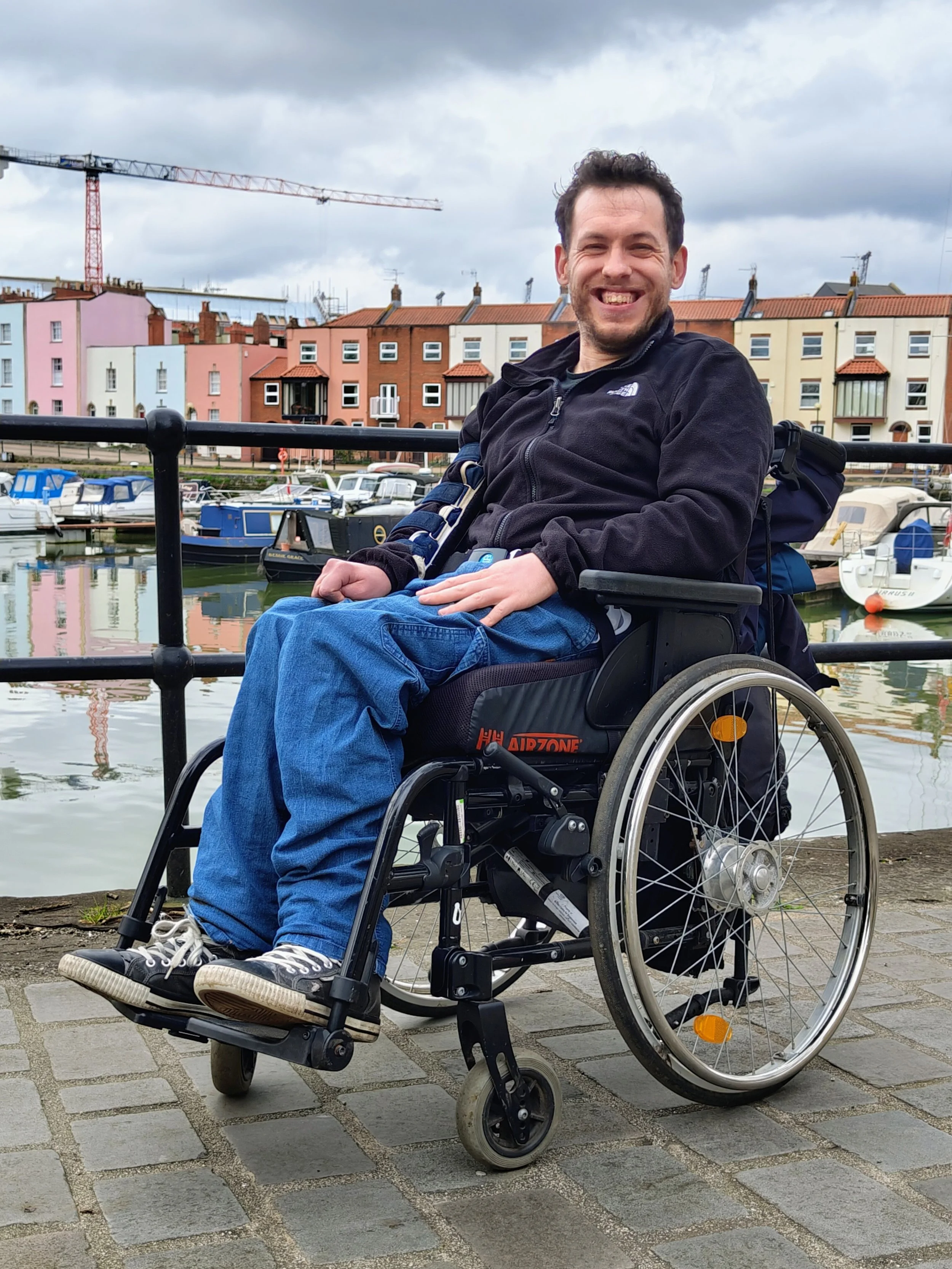 A man sitting in a wheelchair near a marina with boats, colorful buildings, and a cloudy sky in the background.