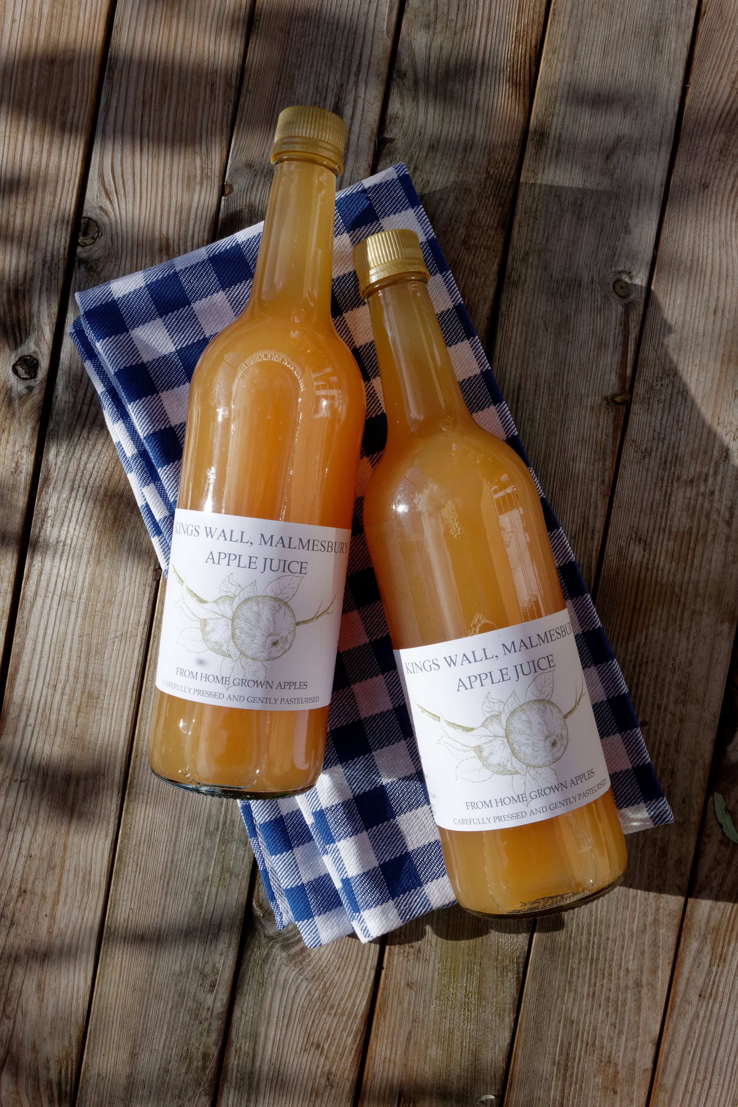 Two bottles of apple juice placed on a wooden surface with a blue and white checkered cloth underneath.