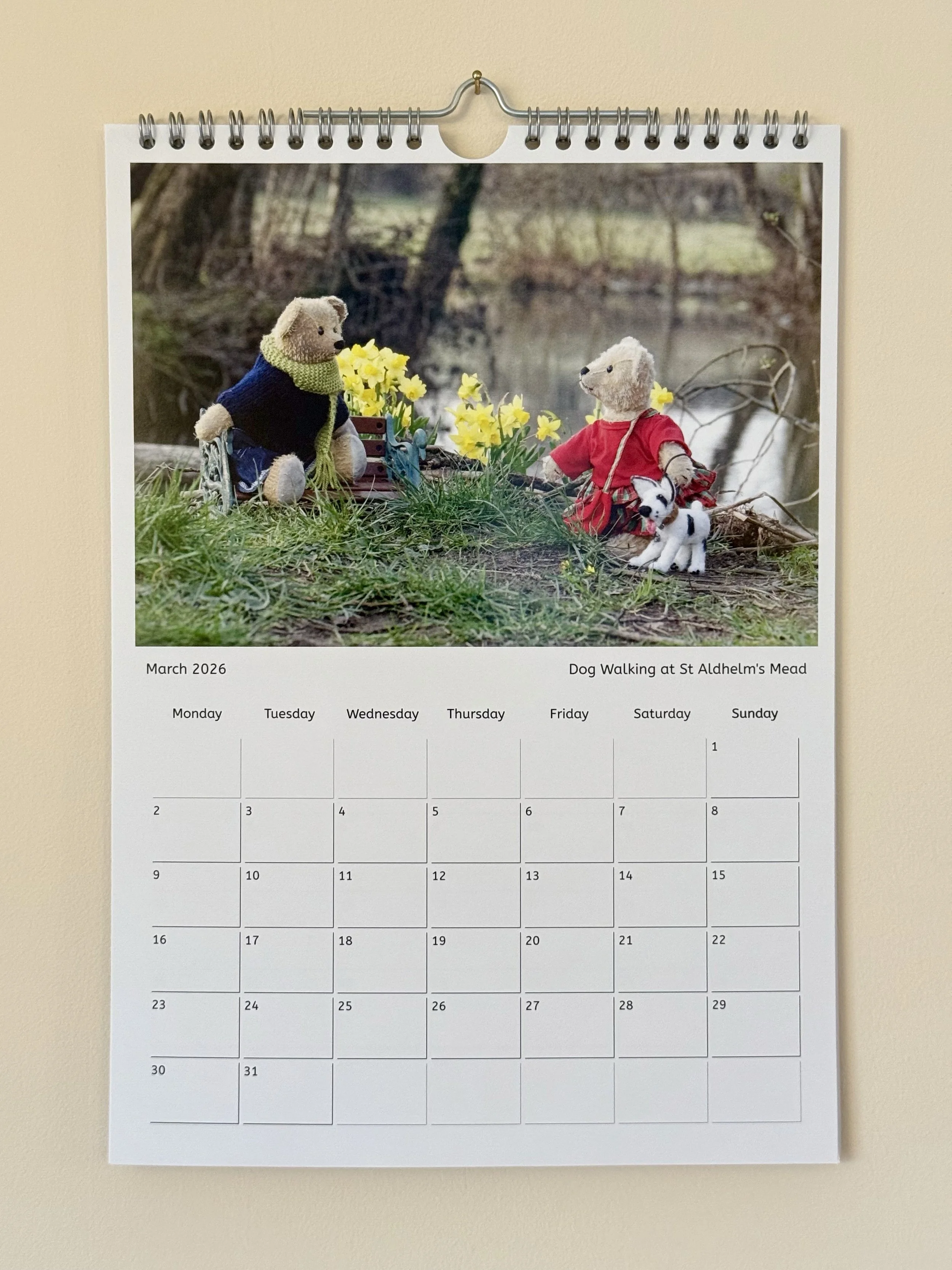 A calendar page for March 2026 featuring a photograph of two teddy bears, one dressed in a navy sweater and scarf, and the other in a red dress with a small dog plush, sitting outdoors near yellow flowers with a blurred natural background.