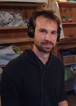 A man with dark hair, a beard, wearing headphones and a black hoodie, standing indoors with wooden shelves and various items in the background.