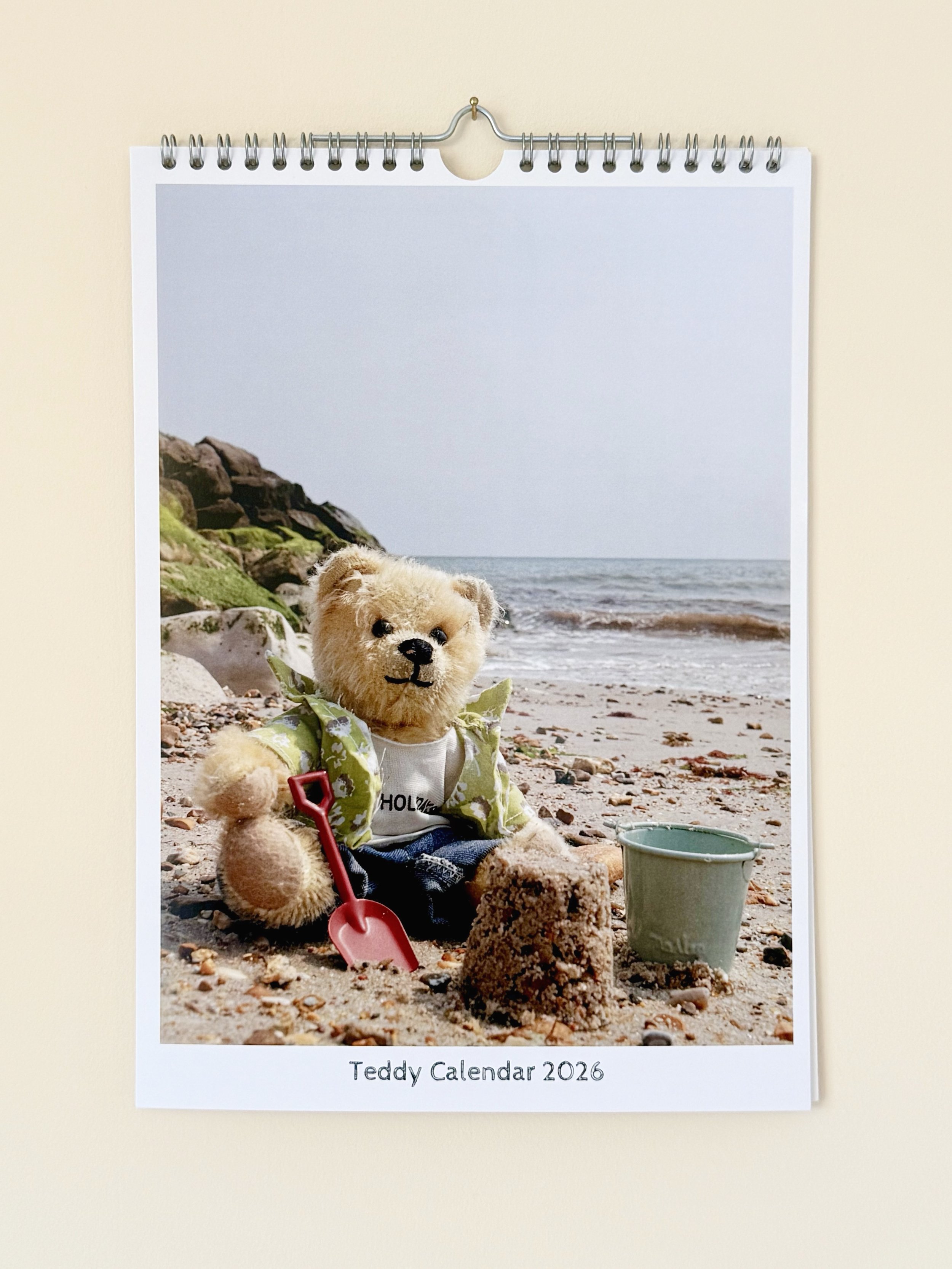 A teddy bear sitting on a sandy beach with rocks, wearing a green shirt and holding a red shovel, next to a small green bucket, with ocean waves in the background, on a 2026 calendar labeled Teddy Calendar.