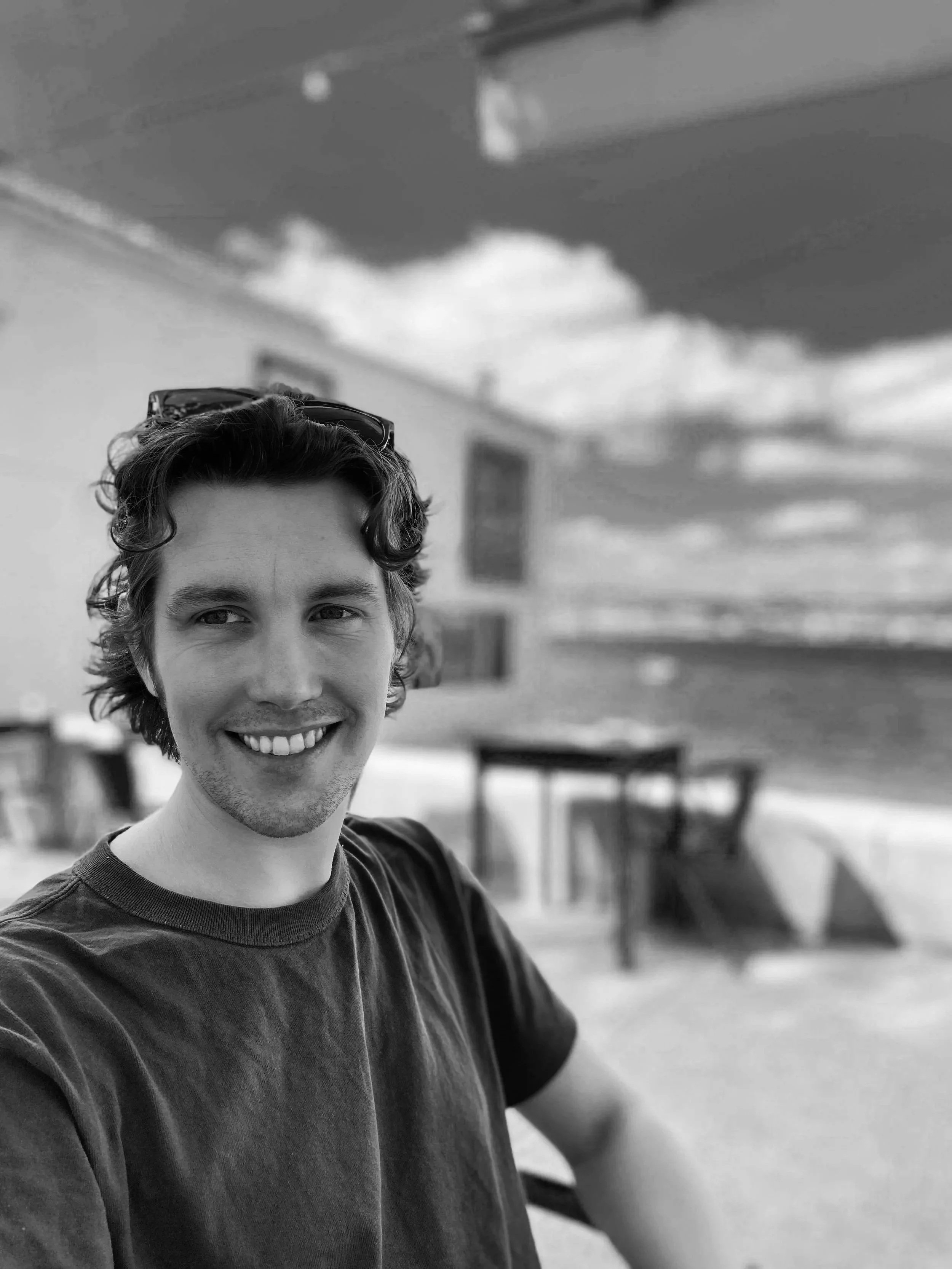 A young man with curly hair smiling and wearing sunglasses on his head, outdoors on a sunny day with a beach and ocean in the background.