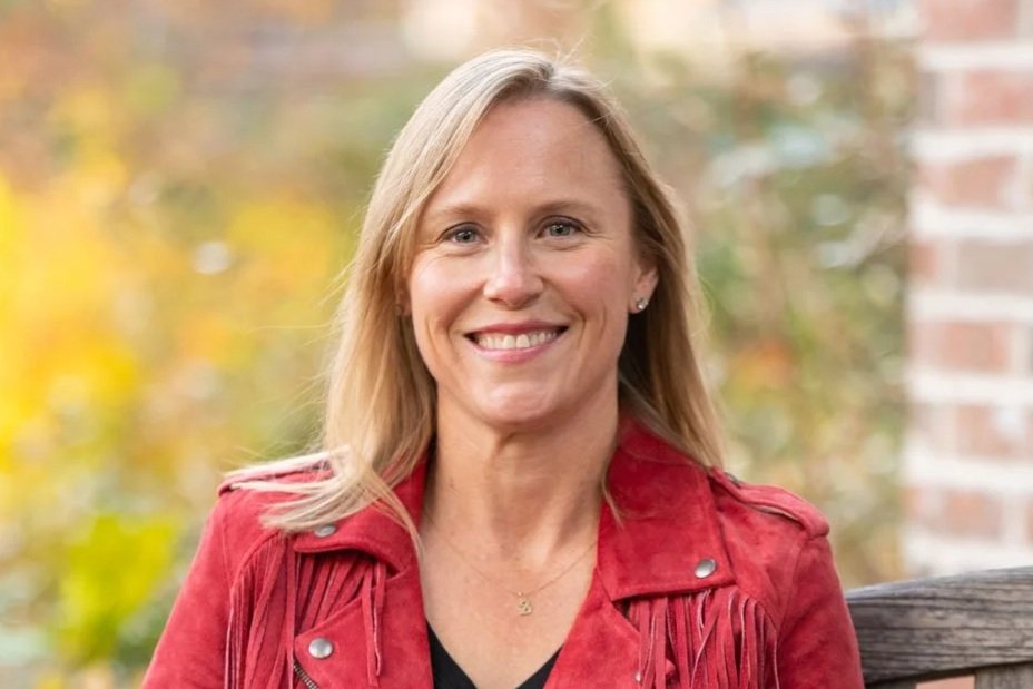 A smiling woman with blonde hair wearing a jacket, sitting outdoors in front of a blurred background with fall foliage.