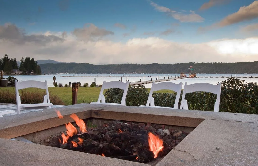 Outdoor fire pit with white chairs around it, overlooking a lake with a dock and boats, trees, and mountains in the background.