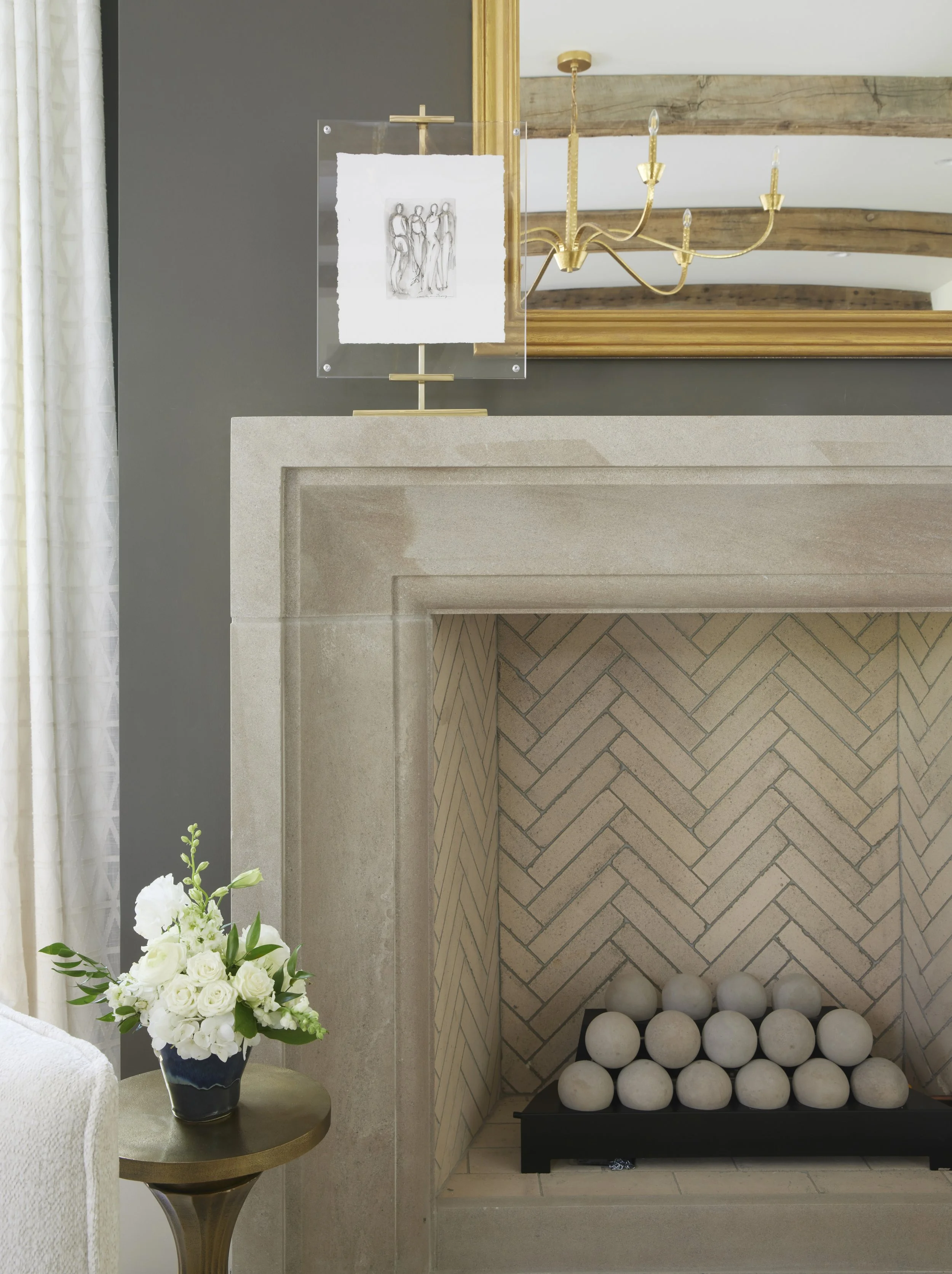 Close-up of a beige fireplace with a black metal grate holding white spheres inside, a bouquet of white flowers on a small round brass table next to it, a gold chandelier, a mirror on the wall, and a piece of wall art above.