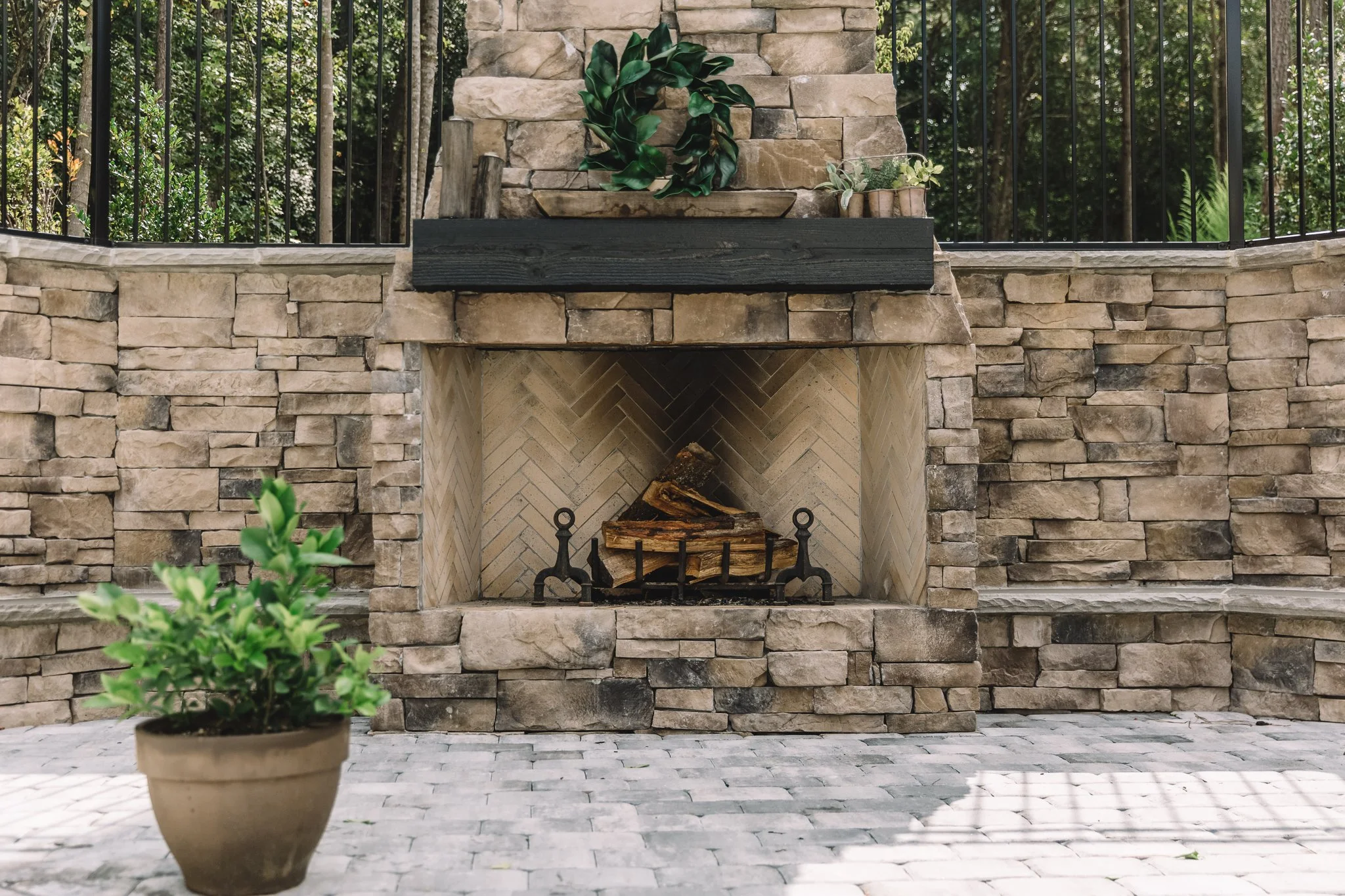 Outdoor fireplace with stacked firewood, surrounded by stone walls and a potted plant in the foreground.