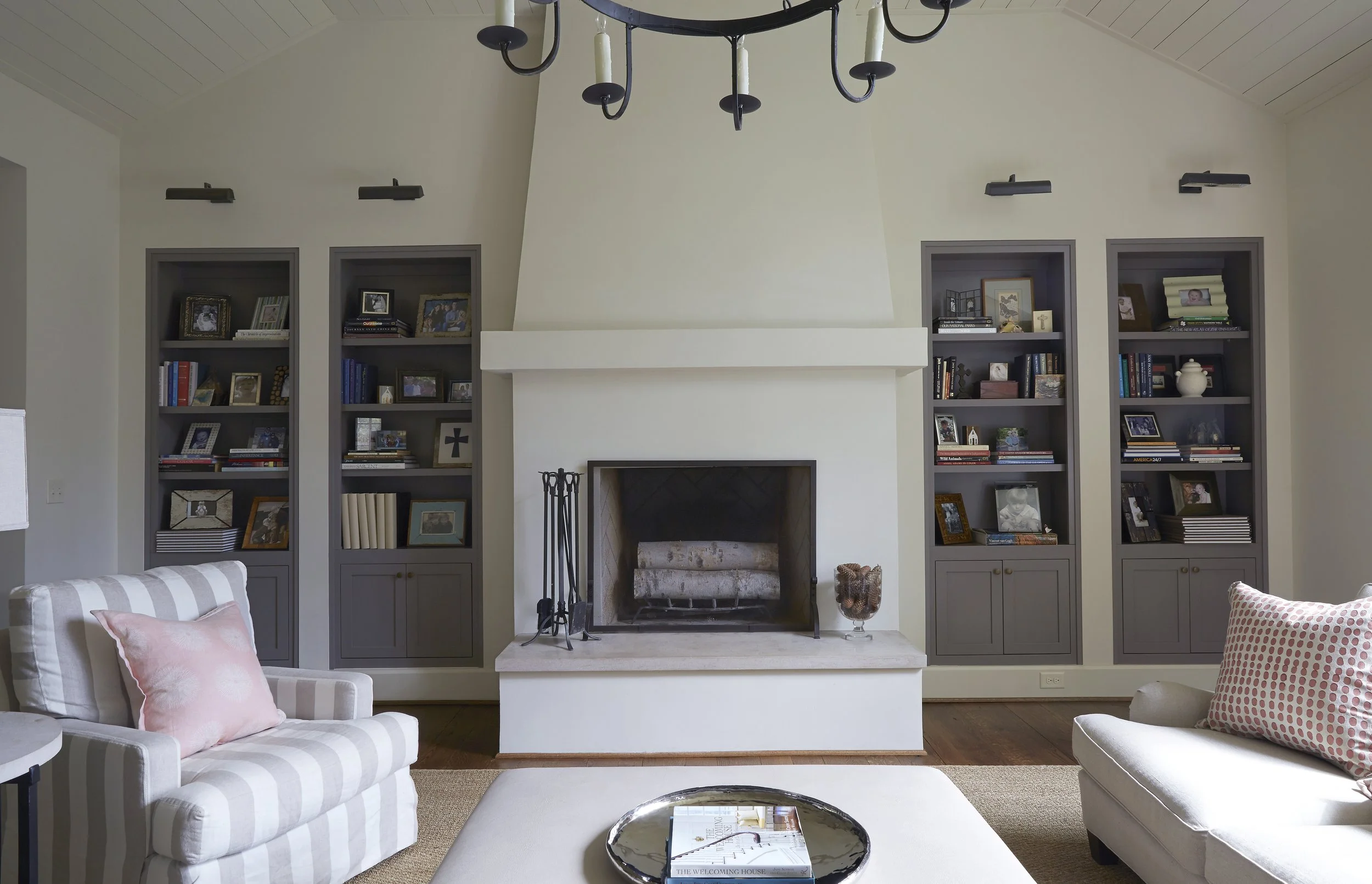 Living room with a white fireplace, gray built-in bookshelves with framed photos and books, two armchairs with pink pillows, a beige ottoman with a tray and books, and a black chandelier.