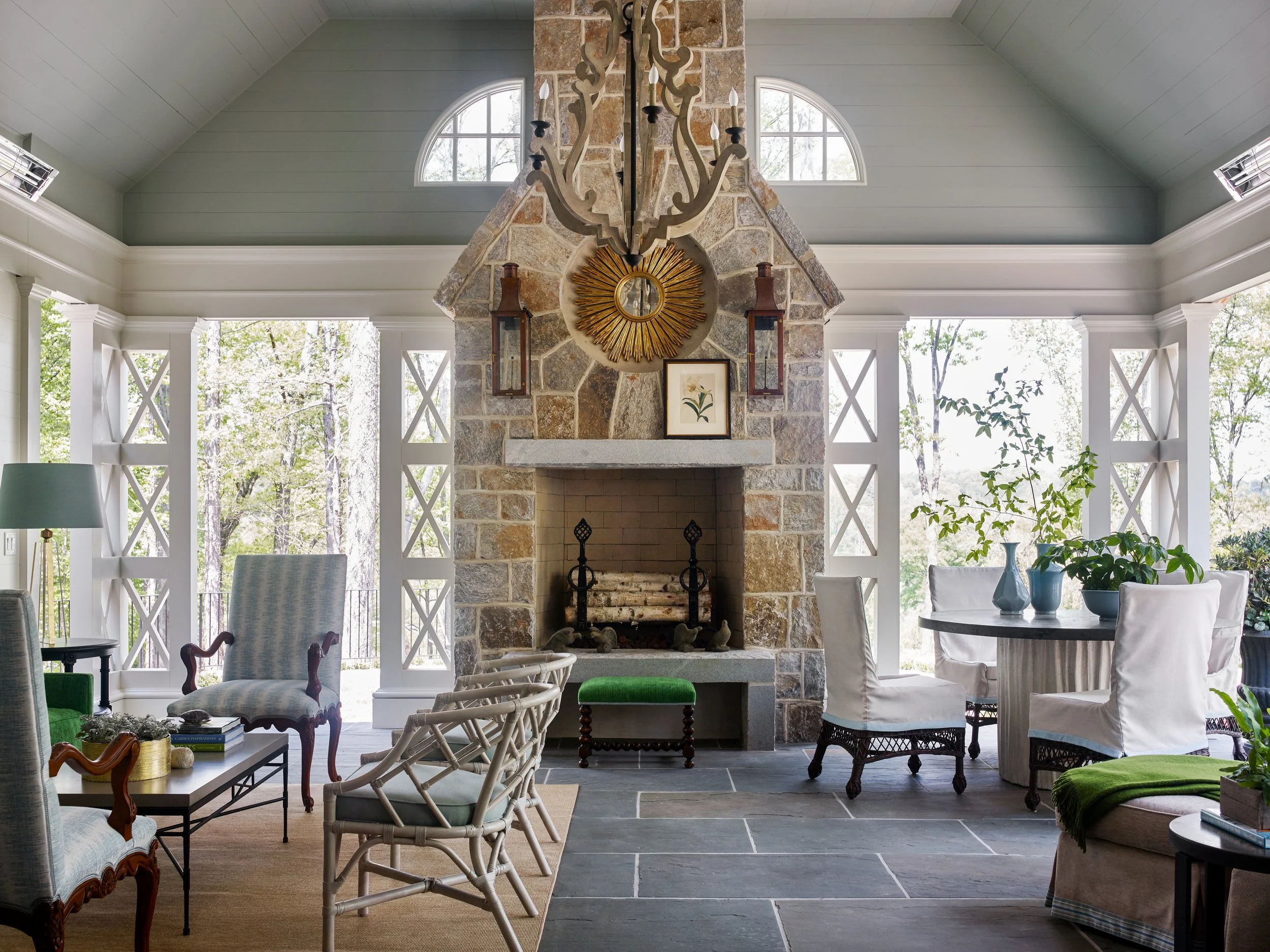 Living room with a stone fireplace, large windows, and deck overlooking a wooded area. Furniture includes armchairs, a dining table with chairs, plants, and decorative lighting fixtures.