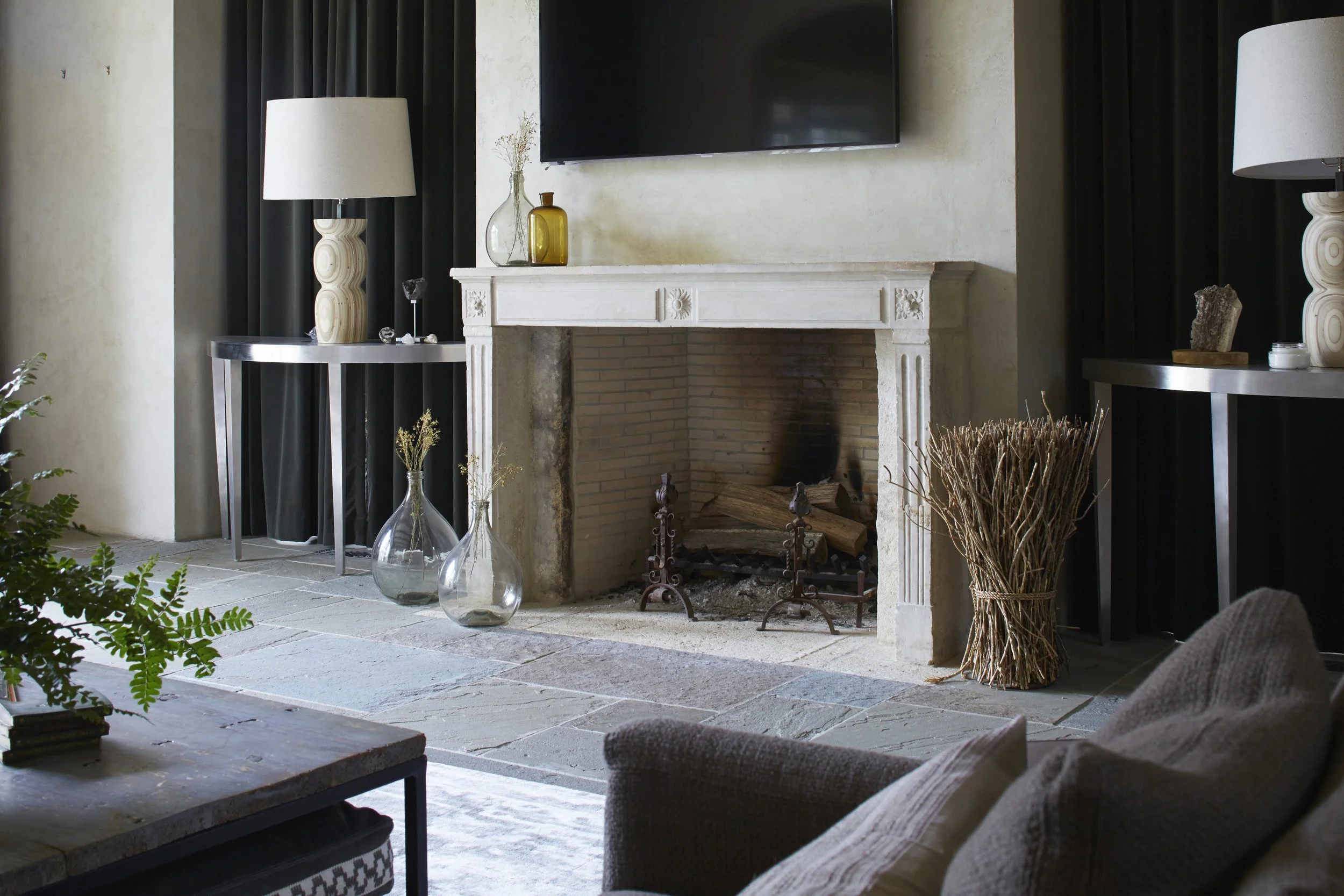 Living room with fireplace, flat-screen TV above, black curtains, two side tables with lamps, decorative vases and objects, green plant, and a beige armchair.