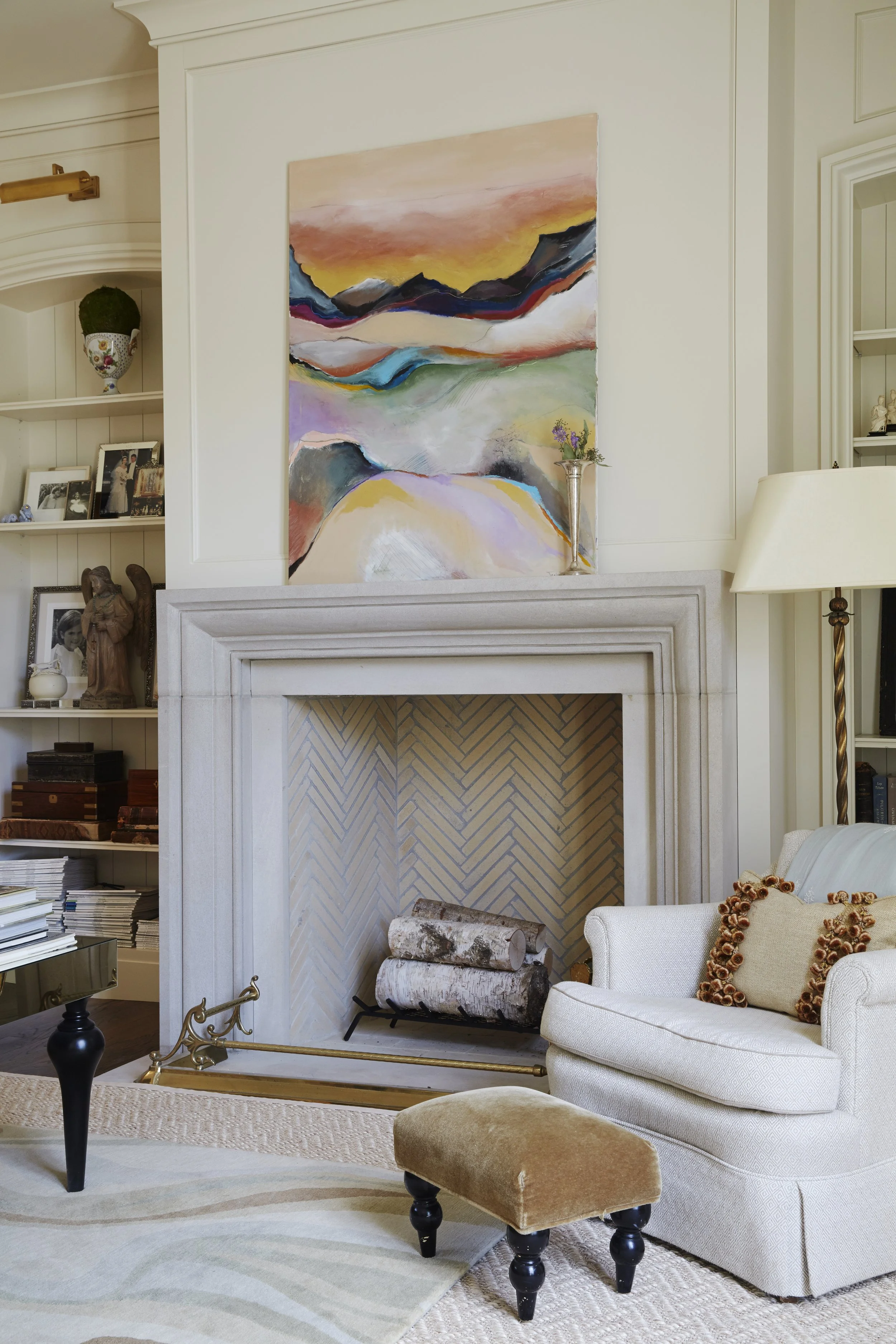 Living room with a stone fireplace, abstract landscape painting above it, upholstered armchair with a decorative pillow, small ottoman, and a lamp on a side table.