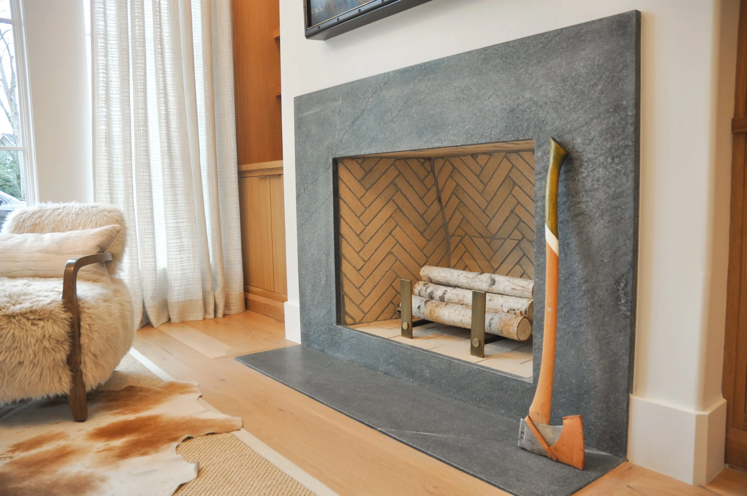 Living room with a modern gray stone fireplace, wooden logs inside, a polished axe leaning against the fireplace, a beige armchair with a furry cover, cream-colored curtains, and natural light coming through a window.