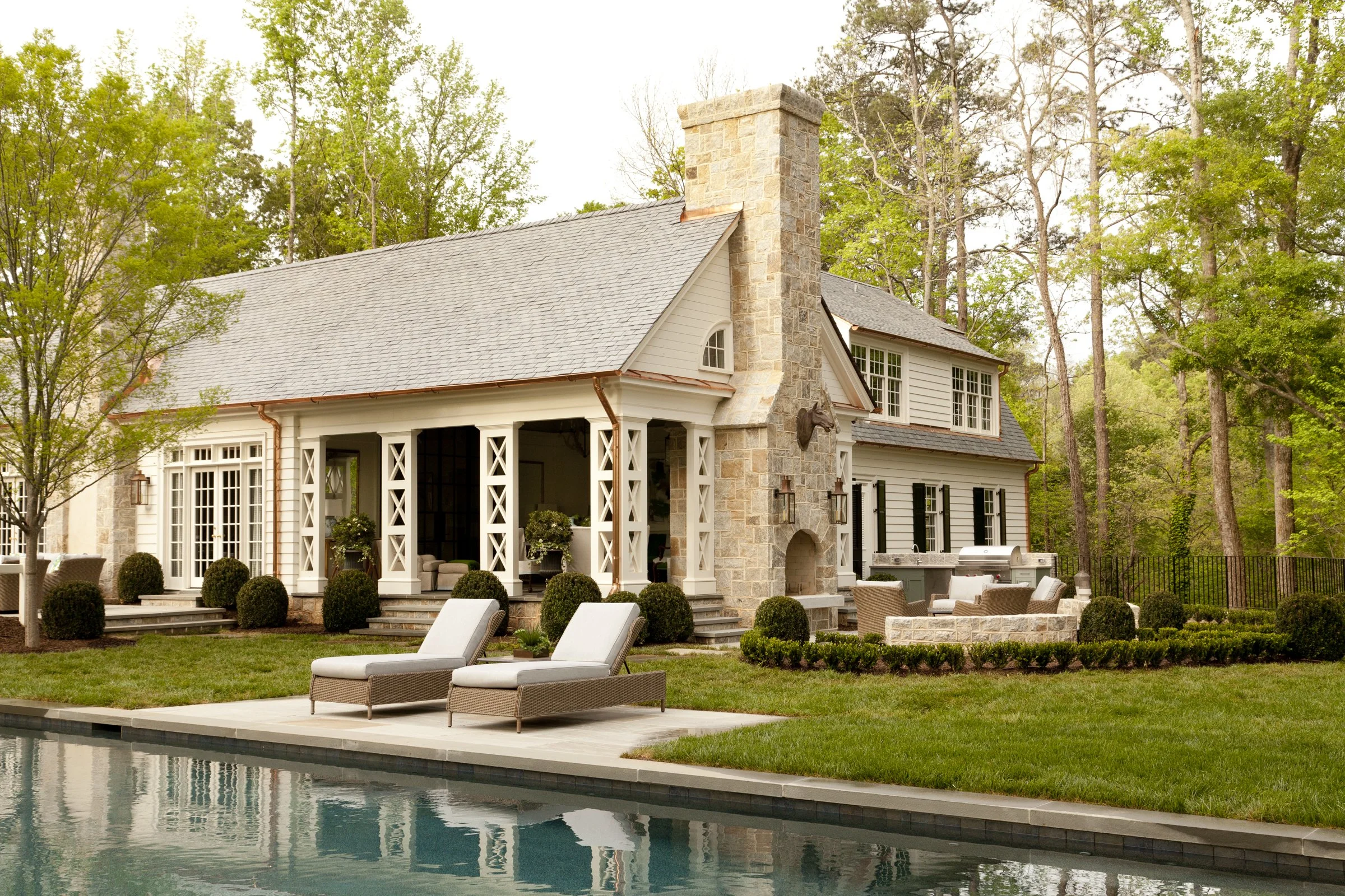 Backyard with swimming pool, lounge chairs, patio area, and house with stone chimney, surrounded by trees.