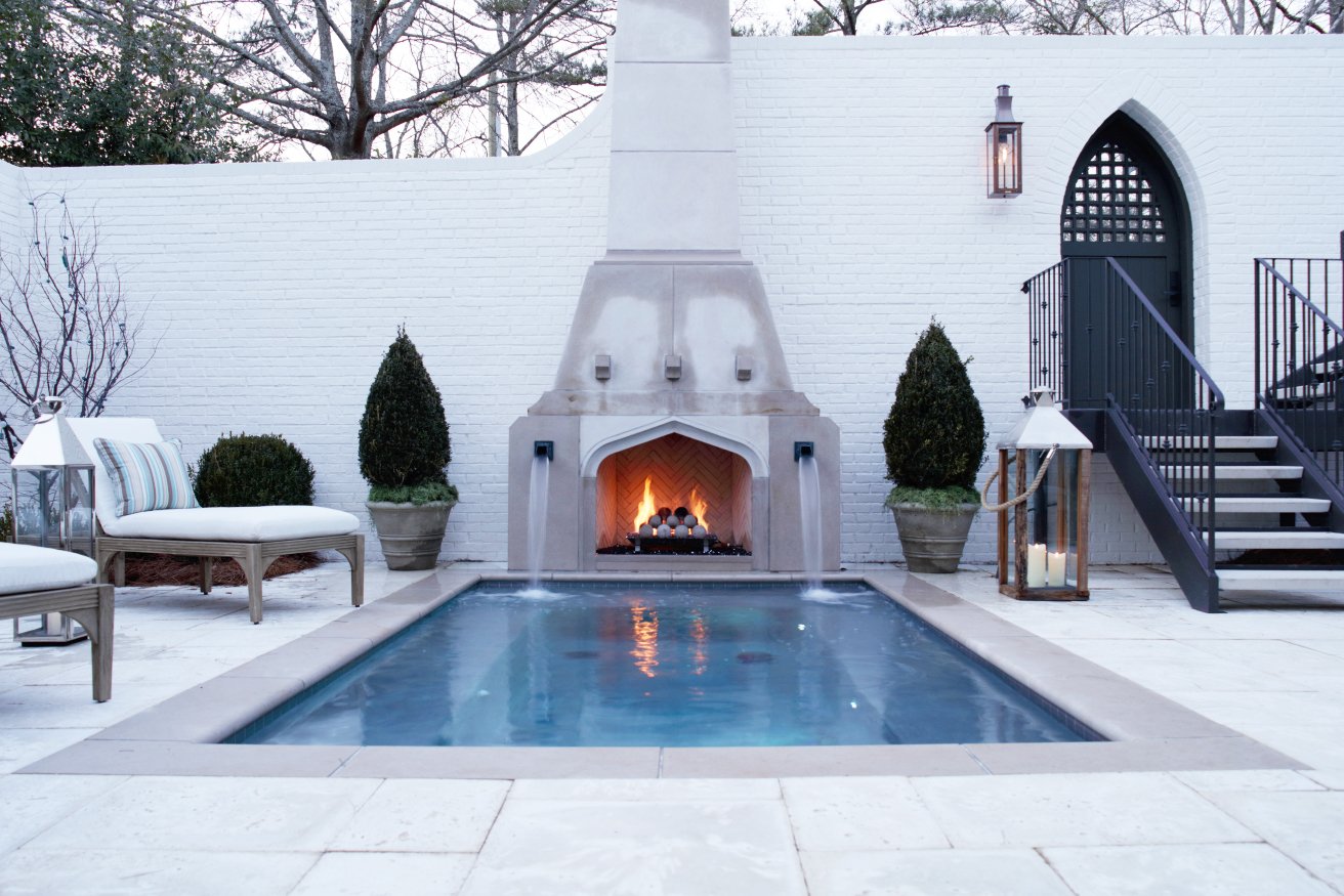 Backyard with a swimming pool, outdoor fireplace with flames, potted shrubs, lounge chairs, lanterns, and a staircase leading to a door.