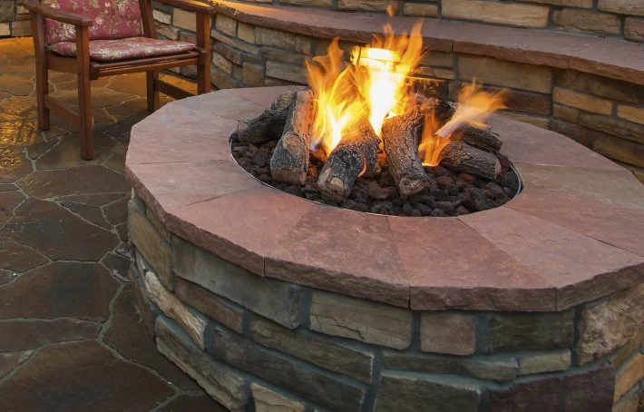 A bubbling fire pit with flames and logs, surrounded by a stone and brick barrier, situated on a stone patio with outdoor furniture.