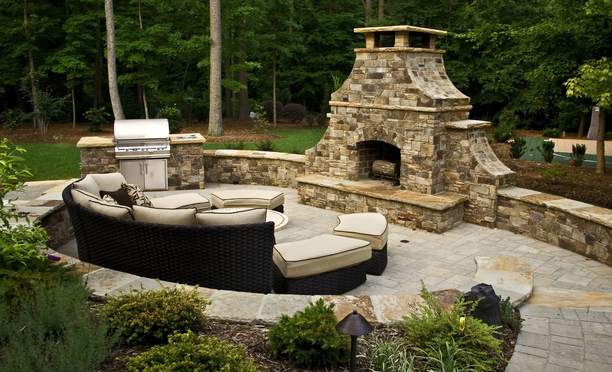 An outdoor patio with a cushioned black wicker sectional sofa arranged in a semi-circle, a stone outdoor fireplace with a chimney, a built-in grill, surrounded by a stone wall, and lush green trees in the background.