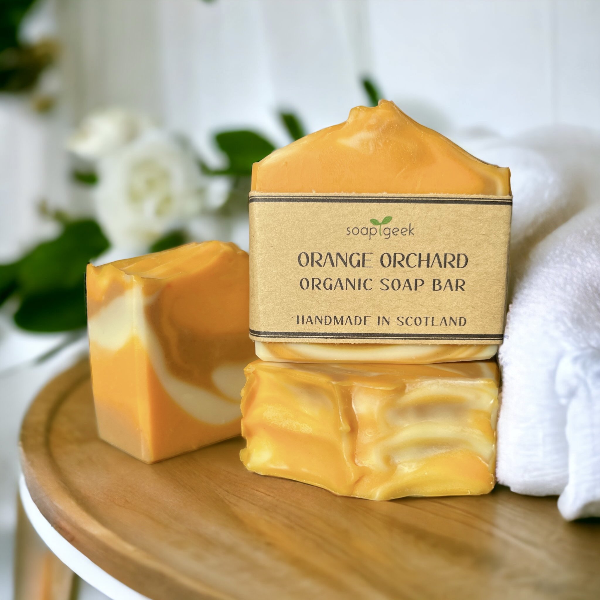 Handmade Orange Orchard soap bars with bright orange and cream swirls, one wrapped in a kraft label, displayed on a wooden surface with white towels and flowers in the background.