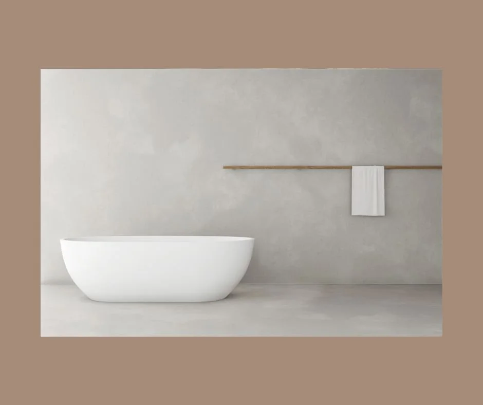 A modern white freestanding bathtub in a minimal bathroom with a textured light grey wall and a wooden towel rail holding a single white towel.