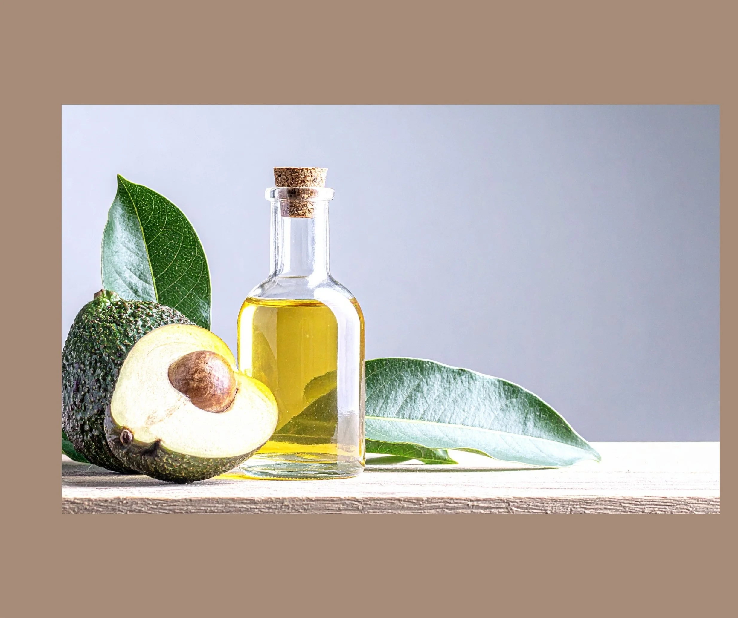 A glass bottle filled with avocado oil next to a halved avocado and green leaves on a light wooden surface.