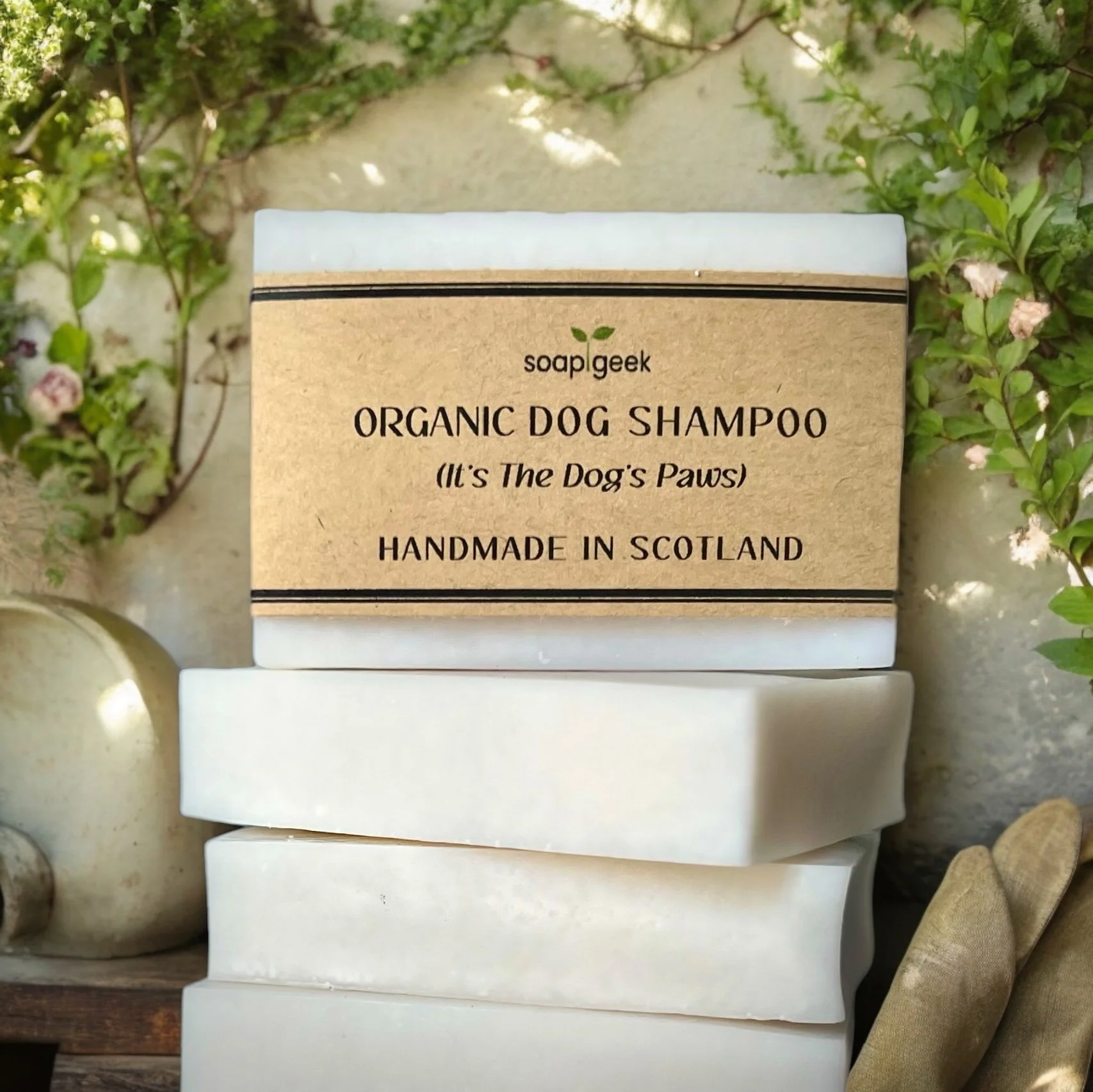 Stack of handmade Organic Dog Shampoo bars with one wrapped in a kraft label, displayed on a rustic wooden surface with green foliage and natural outdoor elements in the background.