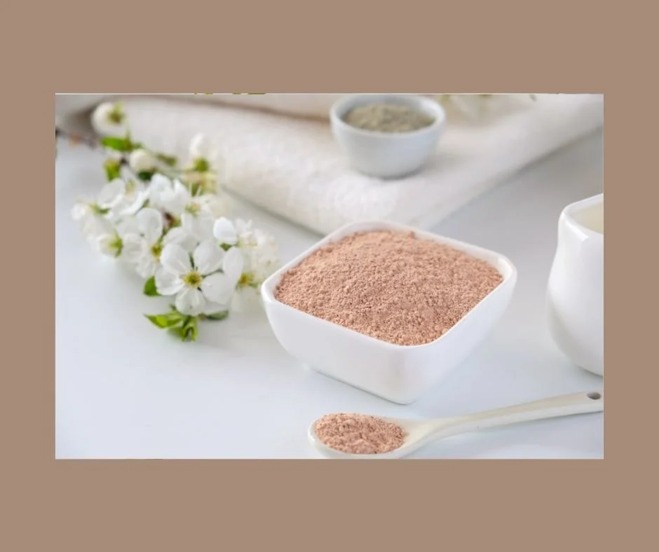 A white bowl filled with pink clay powder on a white surface, with a ceramic spoon holding more clay powder in front, white flowers and folded towels in the background.
