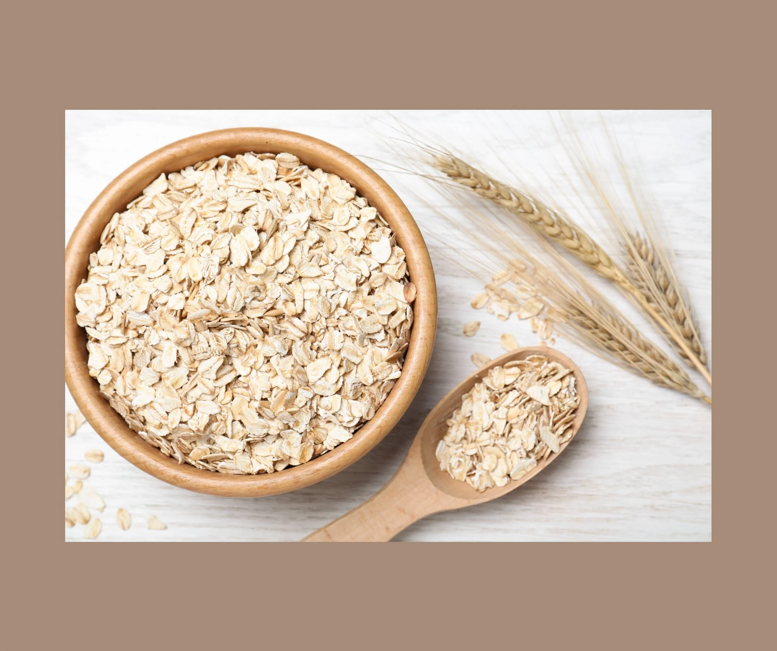 A wooden bowl filled with rolled oats on a light surface, with a wooden spoon holding oats beside it and dried oat stalks in the background.