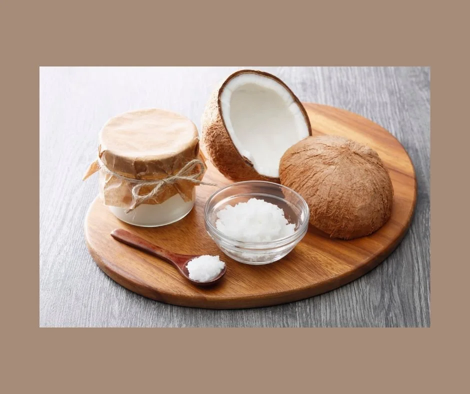 A wooden board holding a glass jar covered with paper and twine, a bowl of coconut oil, a wooden spoon with coconut oil, and a halved coconut.