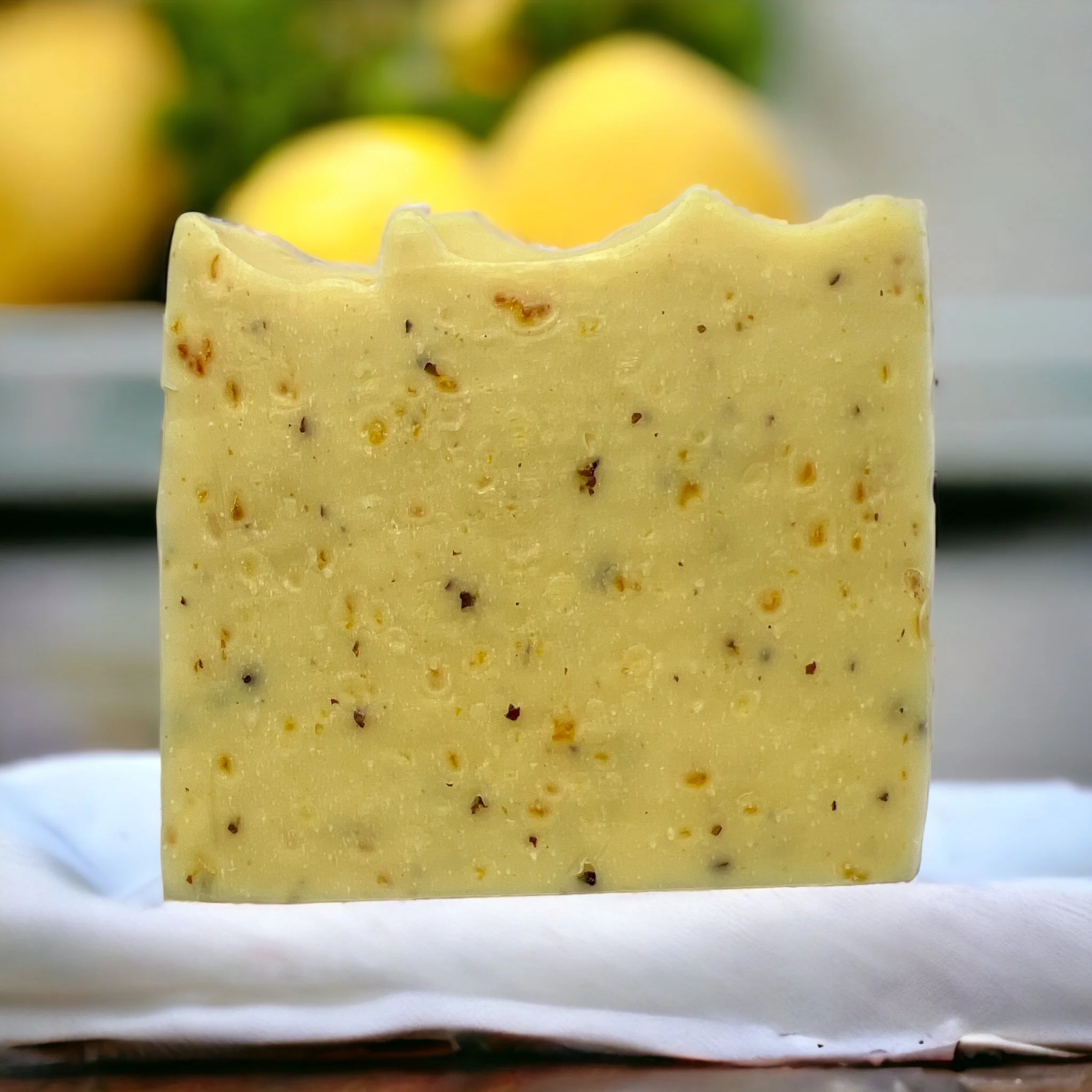 Handmade Lemon & Flaxseed soap bar with a wavy top and flaxseed speckles, standing on a white cloth with whole lemons blurred in the background.