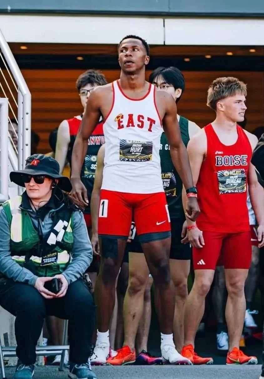 Young male athletes in running gear at a track event, with one wearing a white and red uniform labeled 'EAST' and another in red labeled 'BOISE.' They are preparing for a race.