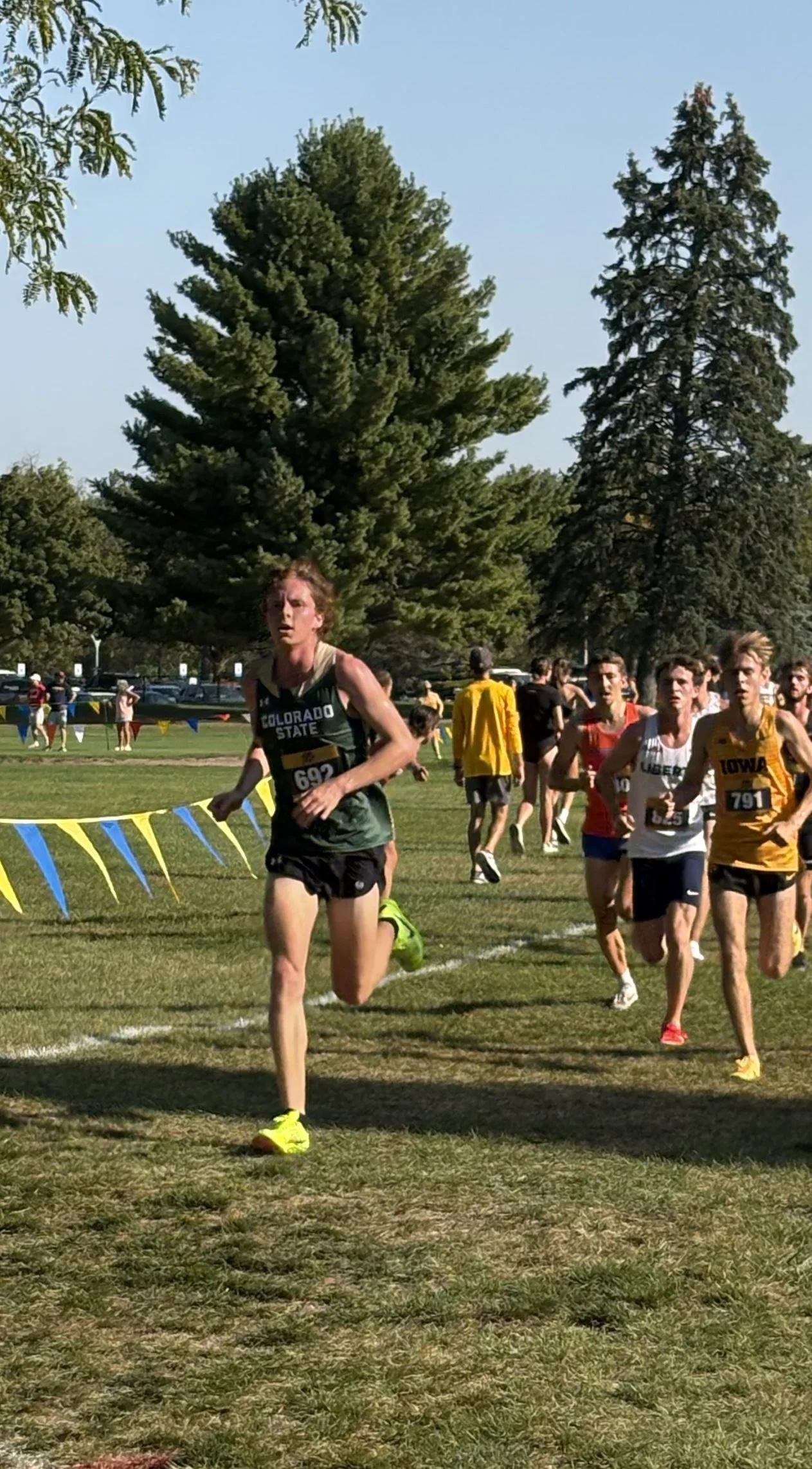 A group of young male runners competing in a cross-country race on a grassy field with trees in the background.