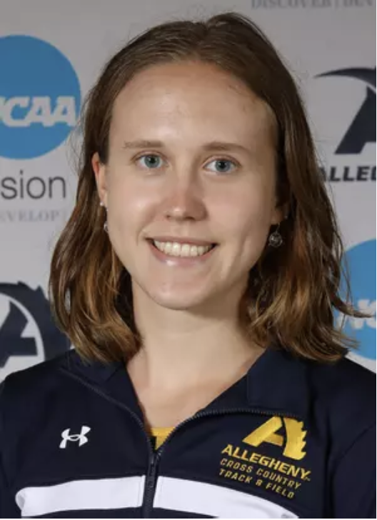 A young woman with shoulder-length red hair and blue eyes, smiling, wearing a navy blue Under Armour jacket with an Allegheny Cross Country Track & Field logo, standing in front of a backdrop with NCAA logos.