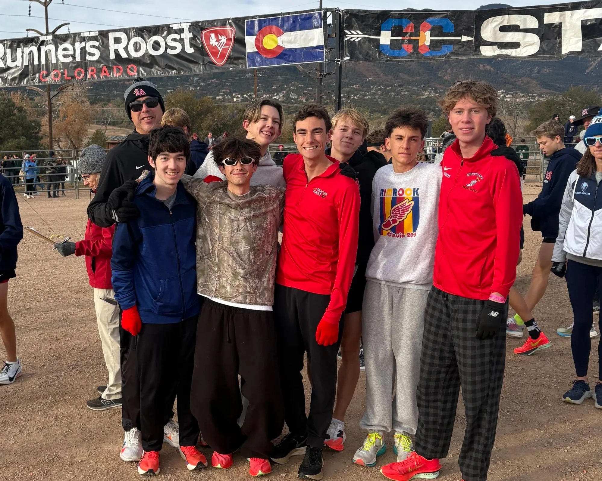 A group of boys and a man at a cross country race in Colorado, standing together and smiling in front of a festival arch and mountains in the background.