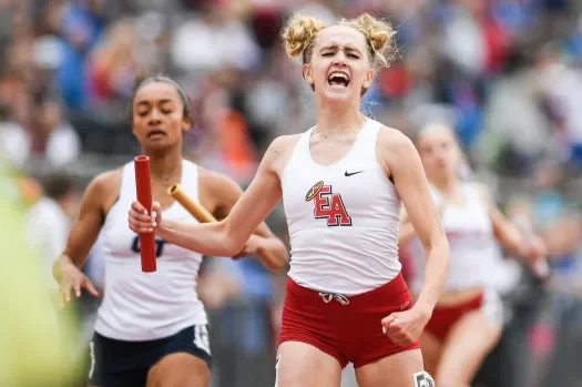 A female athlete celebrating after a relay race, holding a baton and smiling, with other runners and spectators in the background.