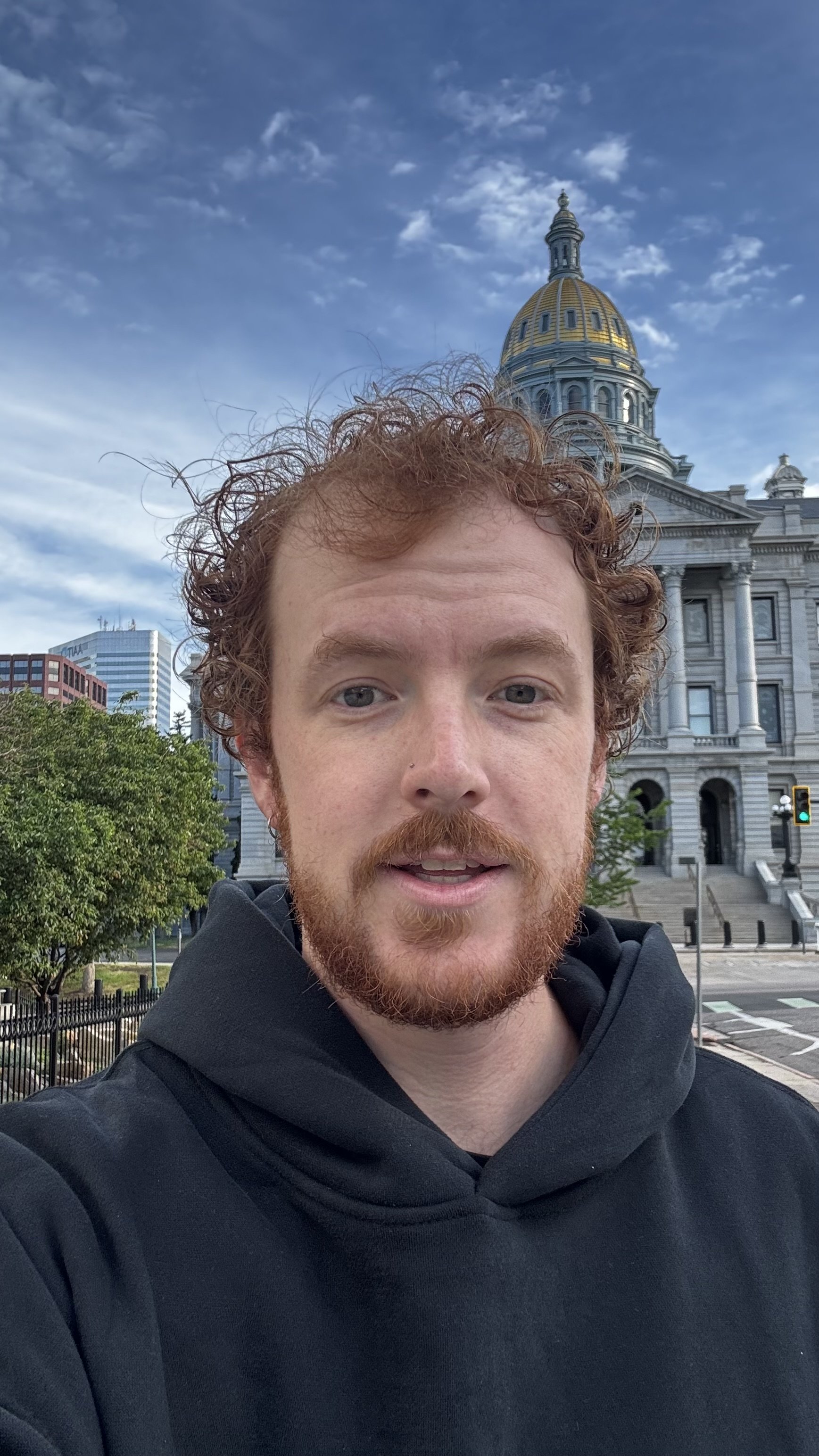 A young man with red curly hair and a beard taking a selfie in front of a historic government building with a gold-domed roof, with modern skyscrapers in the background.