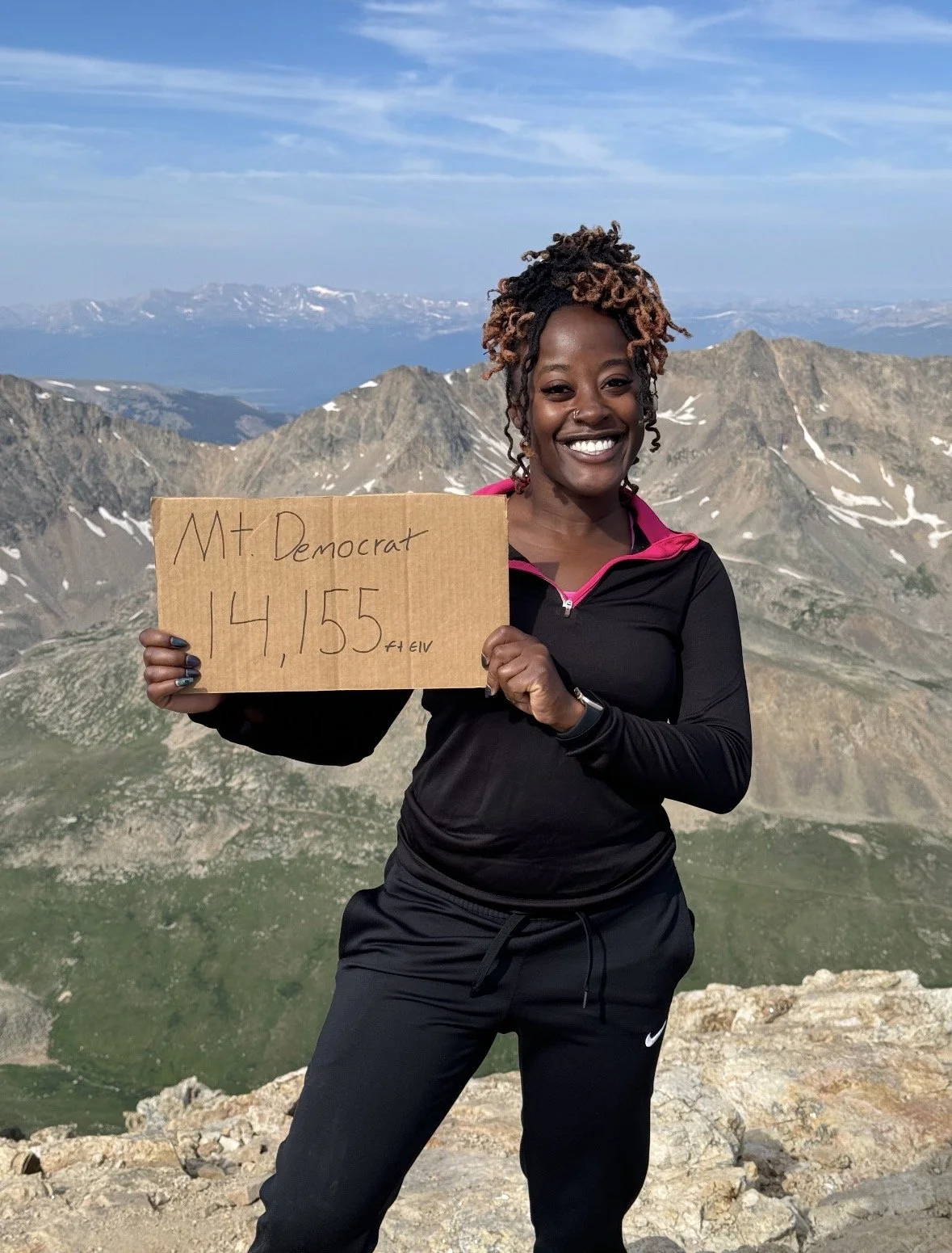 A woman with curly hair holding a cardboard sign that says 'Mt. Democrat 14,155 ft' standing on a mountain with a scenic view of snow-capped mountains and blue sky in the background.
