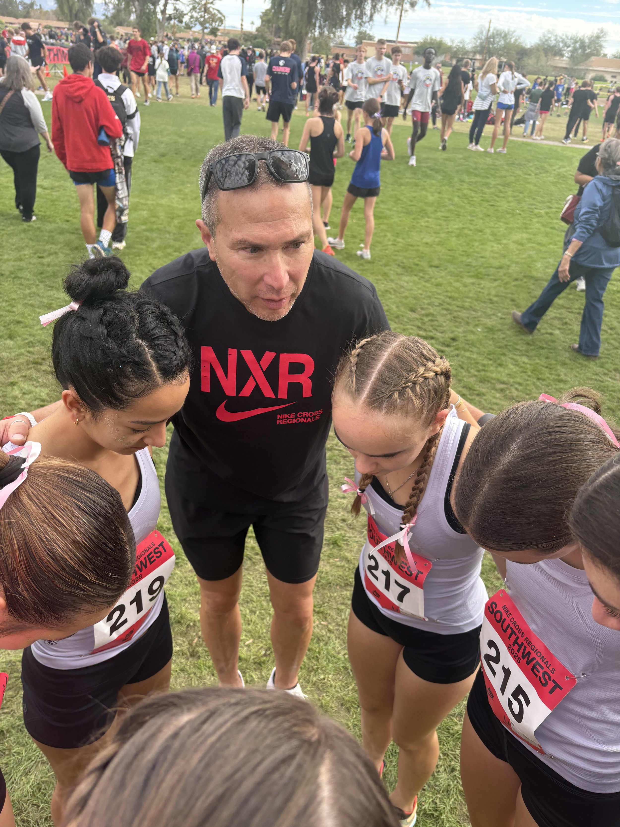 Man coaching young female runners in a huddle at a cross-country meet, with other athletes and spectators in the background.
