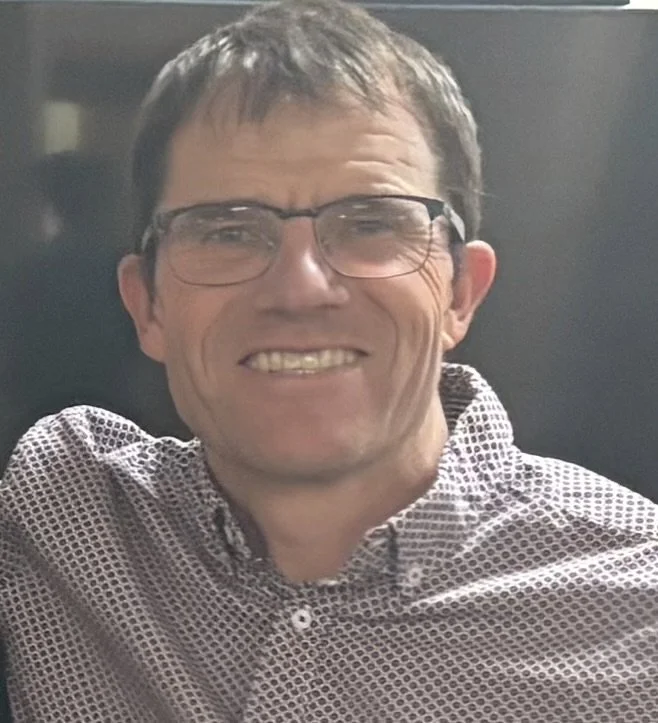 A man with glasses and short brown hair smiling, wearing a patterned button-up shirt.